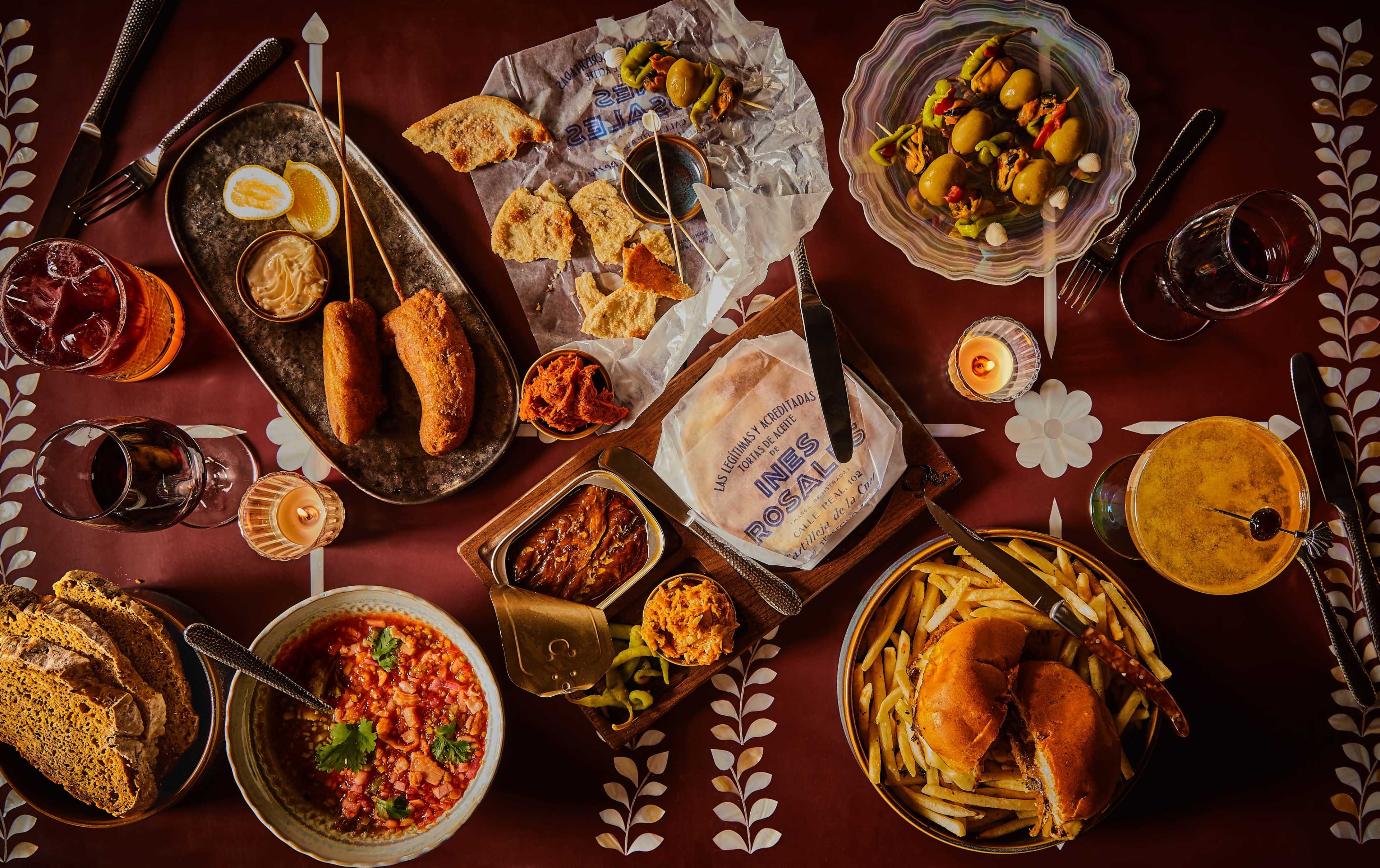 The image showcases a table filled with various dishes, including fried foods, salads, breads, and beverages, arranged on a richly colored tabletop.