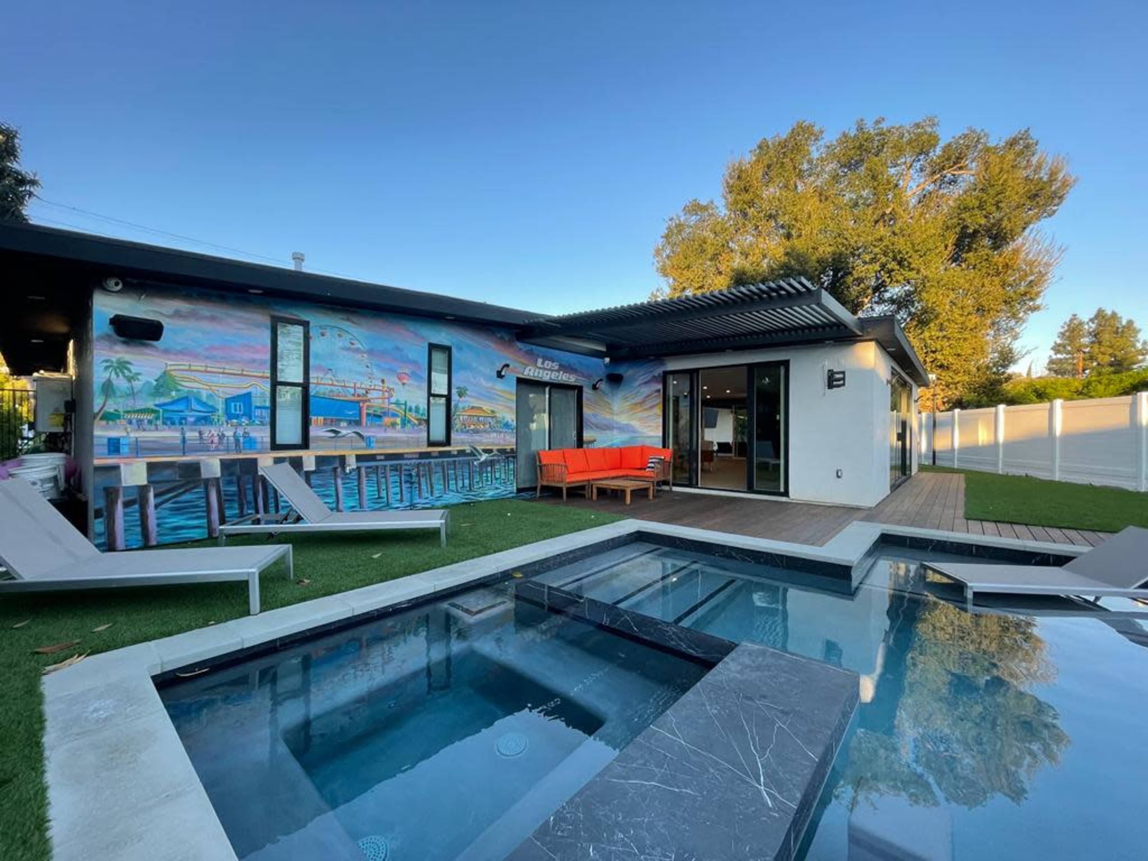 A modern house features a colorful mural on its exterior, an outdoor seating area with an orange couch, and a small pool beside a wooden deck.