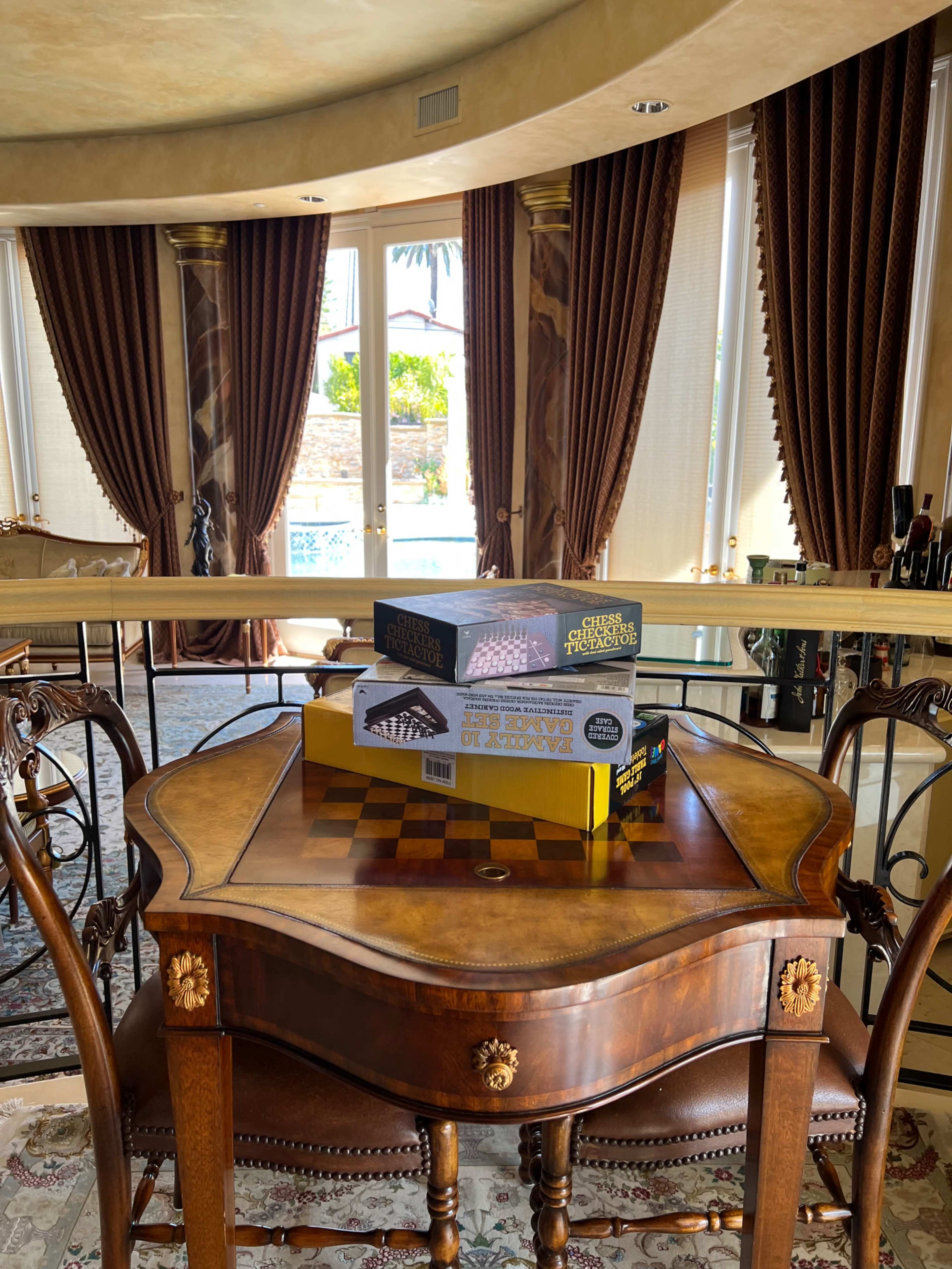 A chess set and a board game box are displayed on a polished wooden table in a room with large windows and elegant curtains.