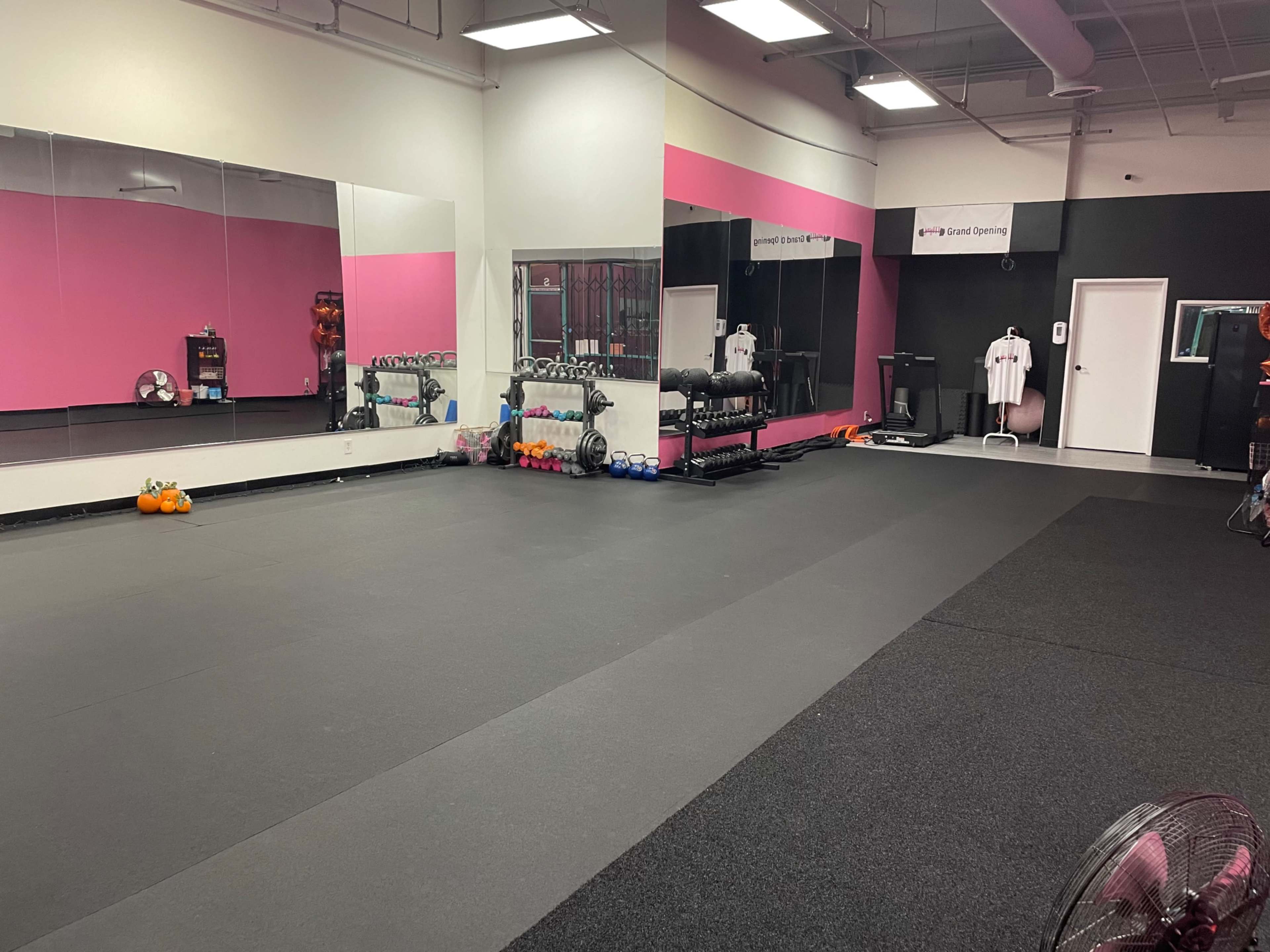 The image shows a gym interior with pink and black walls, featuring a mirrored wall, various exercise equipment, and free weights on the floor.