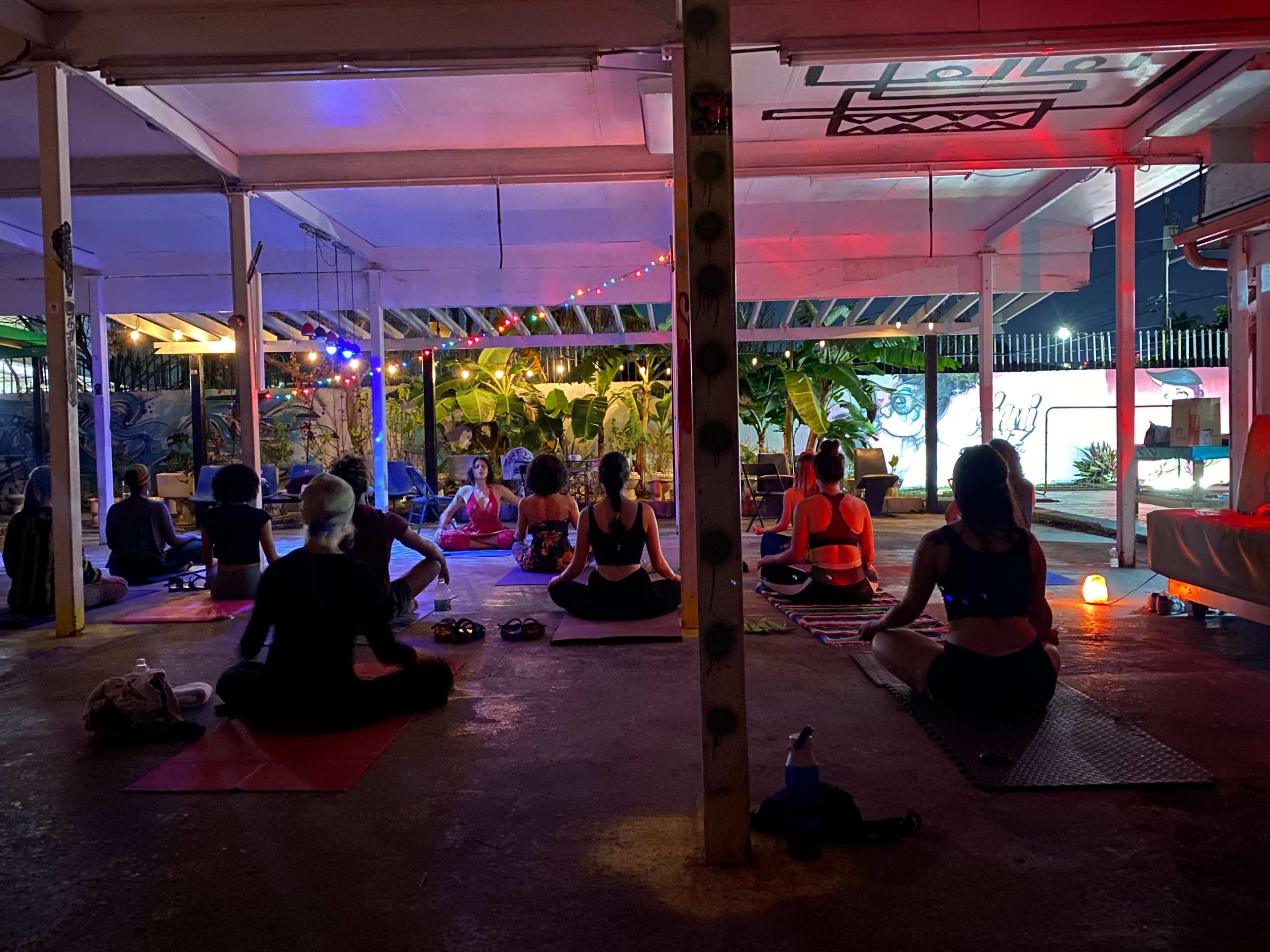 A group of people practice yoga in a dimly lit outdoor space decorated with colorful lights and surrounded by greenery.