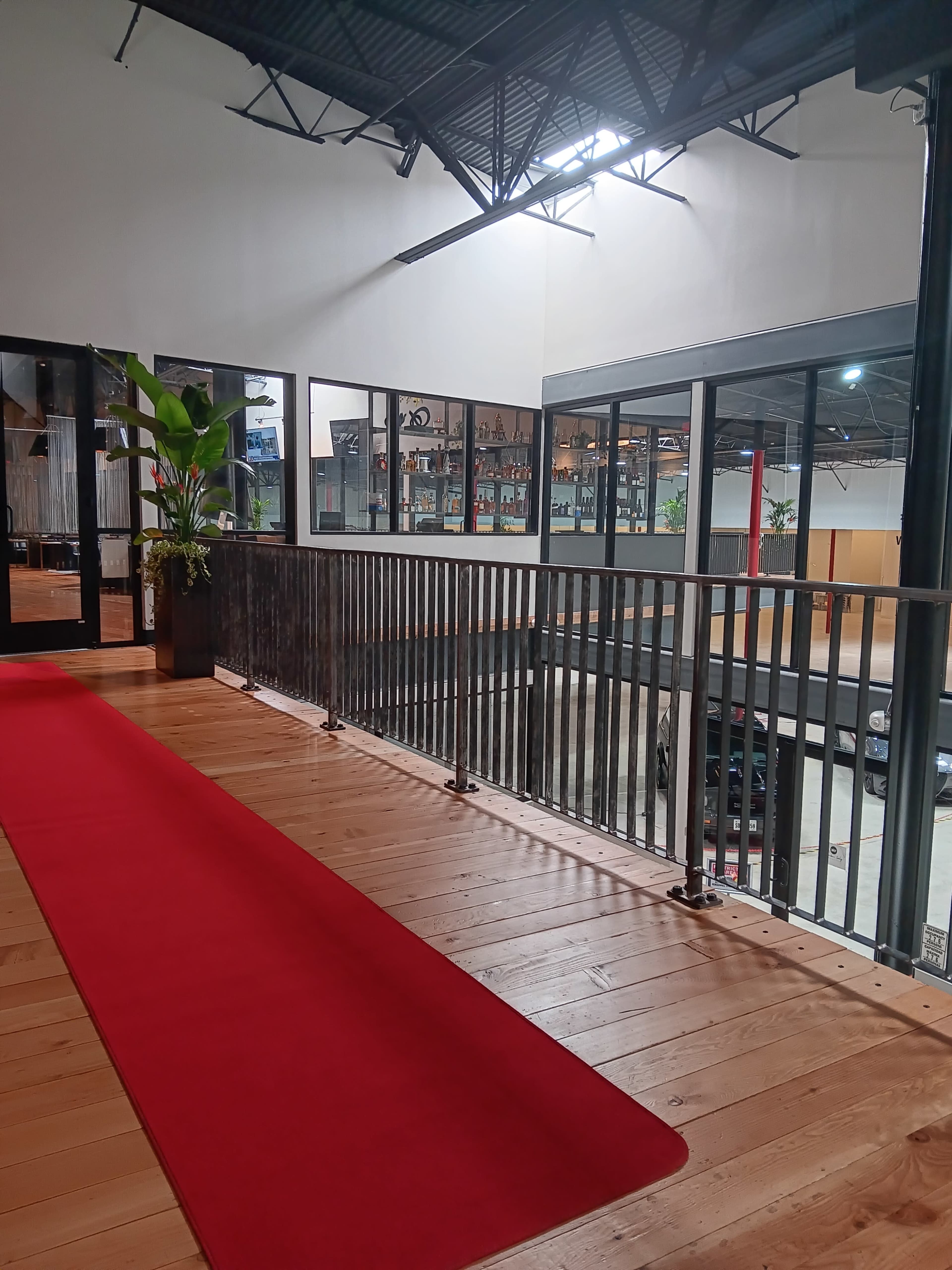 The image shows a wooden interior with a red carpet leading towards a balcony overlooking a parking area, framed by large windows and modern decor.