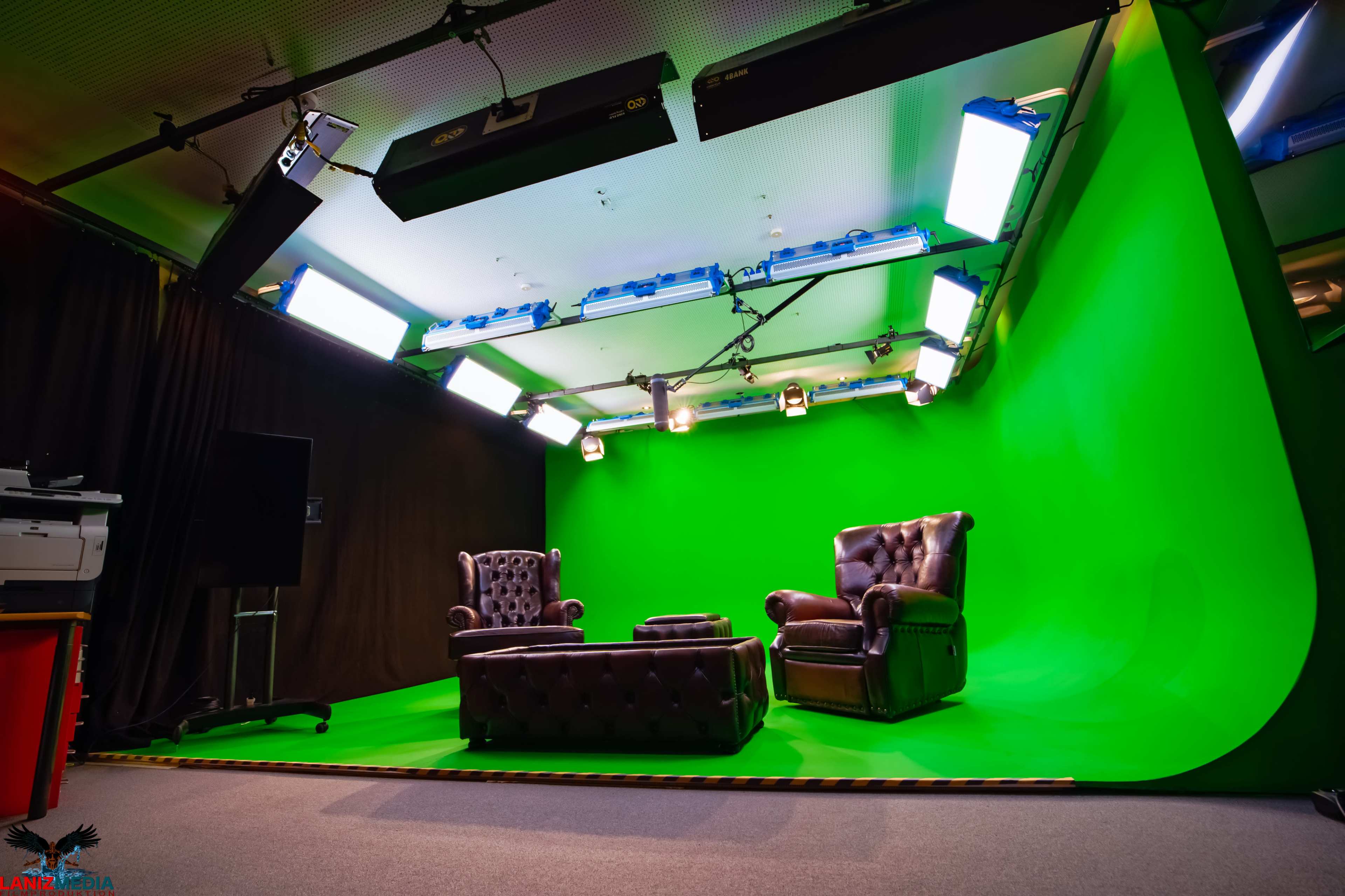 The image shows a studio setup featuring two armchairs and a coffee table in front of a green screen, with lights mounted above.