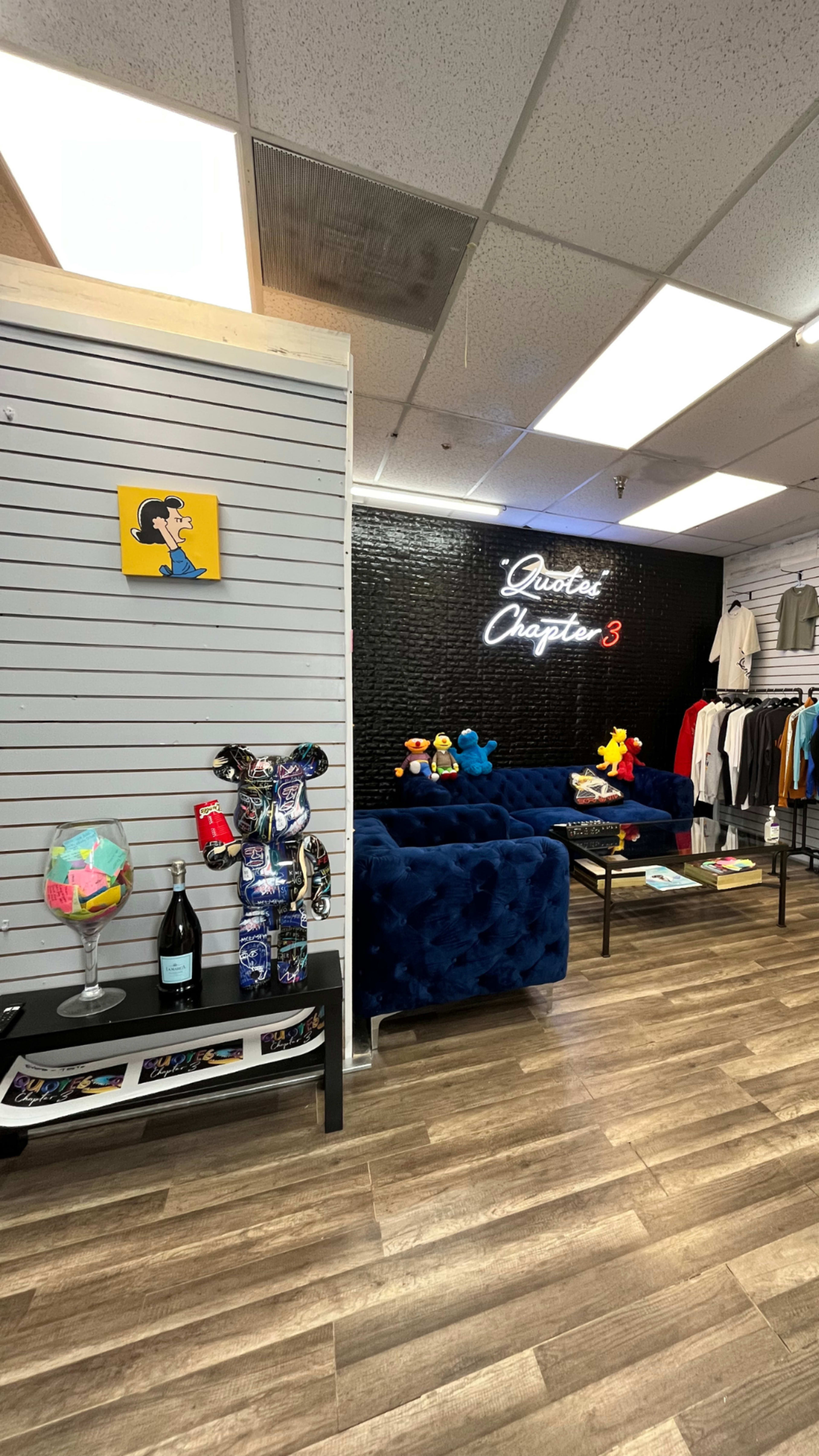A retail store interior featuring a blue velvet sofa, a display table with decorative items, and clothing racks along the walls.