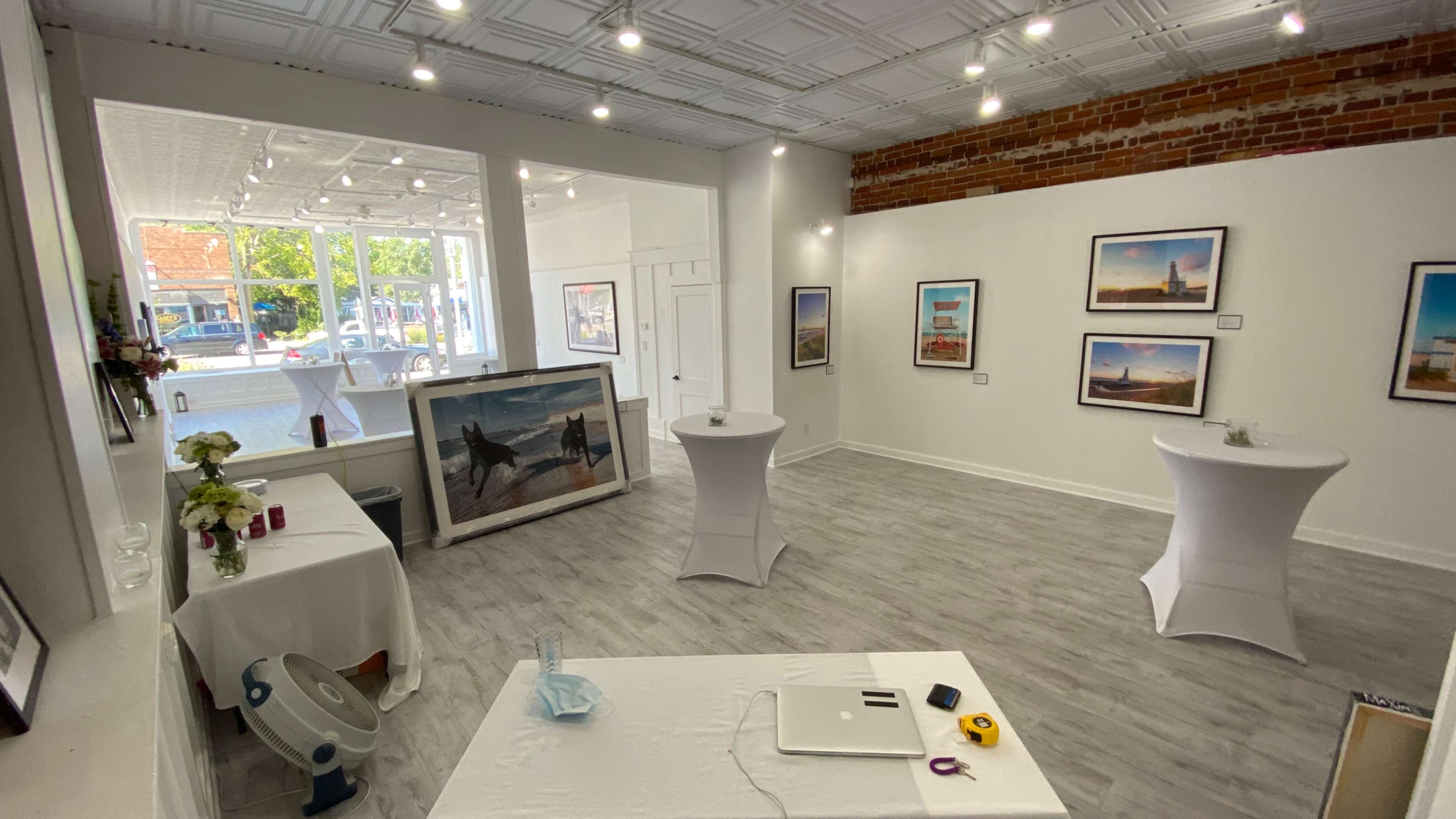 The image shows an art gallery featuring white tables, framed photographs on the walls, and a view of windows letting in natural light.