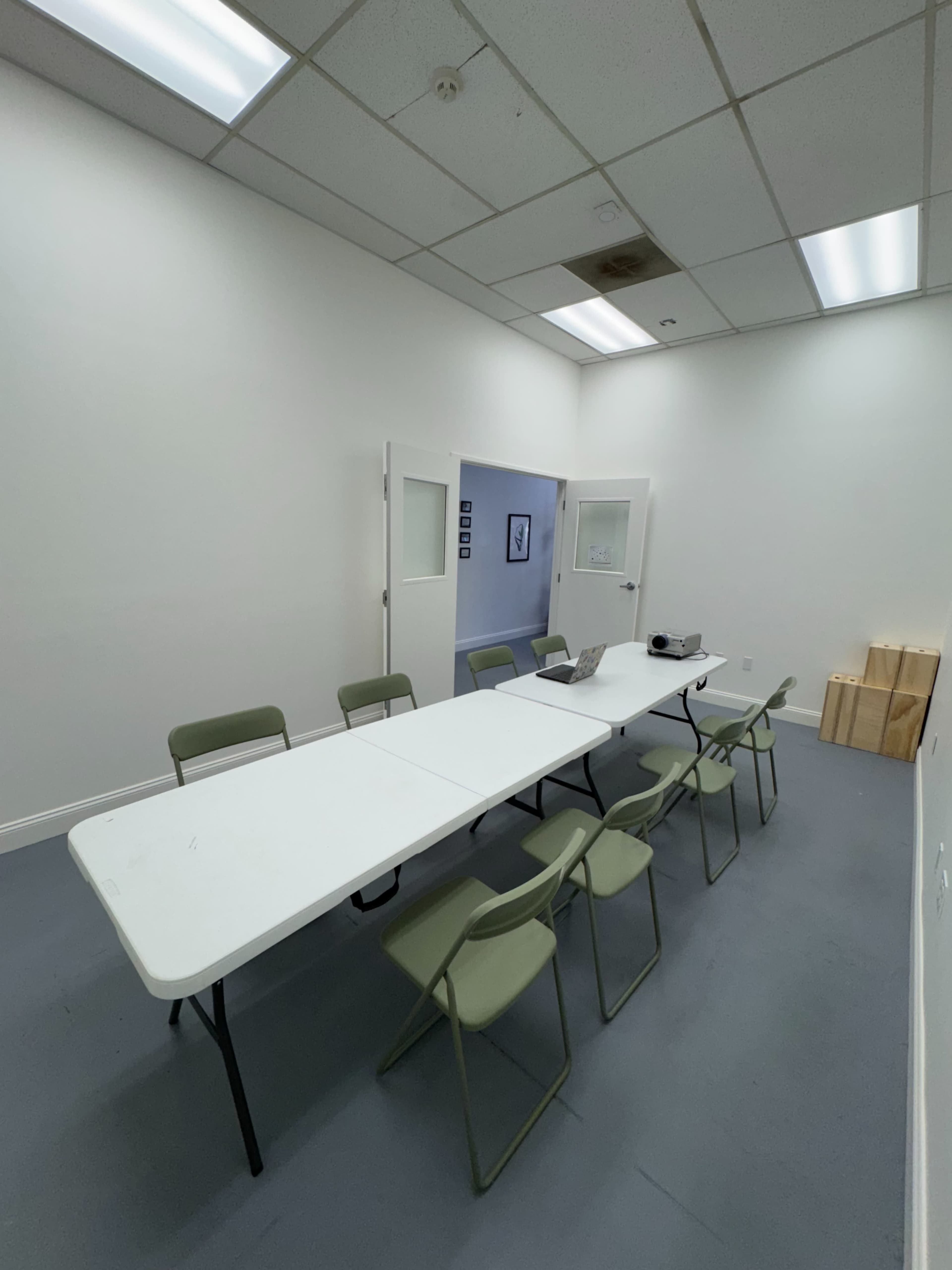 A long table with several chairs is set up in a sparsely furnished meeting room with white walls and a gray floor.
