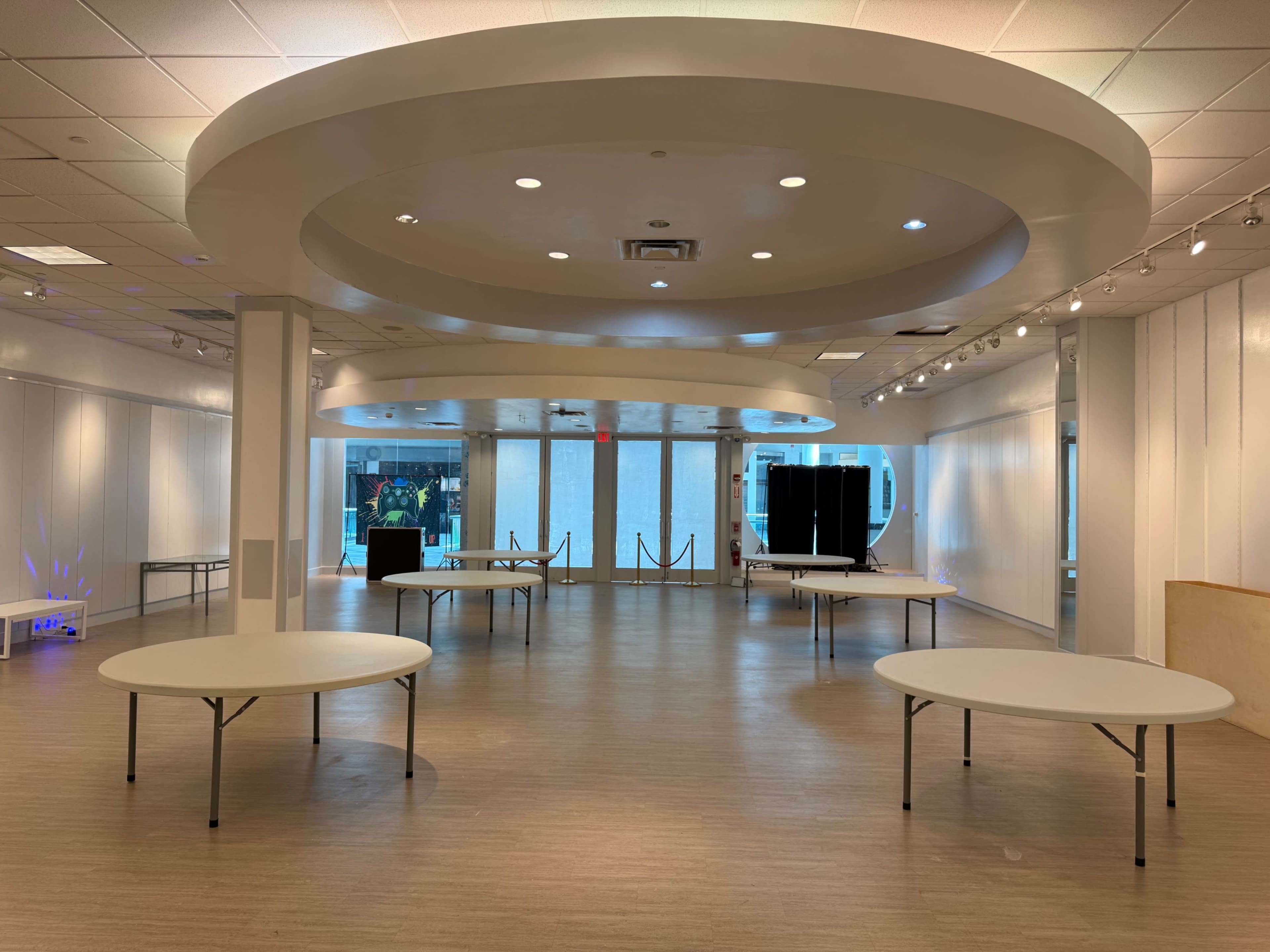 The image shows an empty, well-lit room with several round tables arranged in a spacious, open layout.