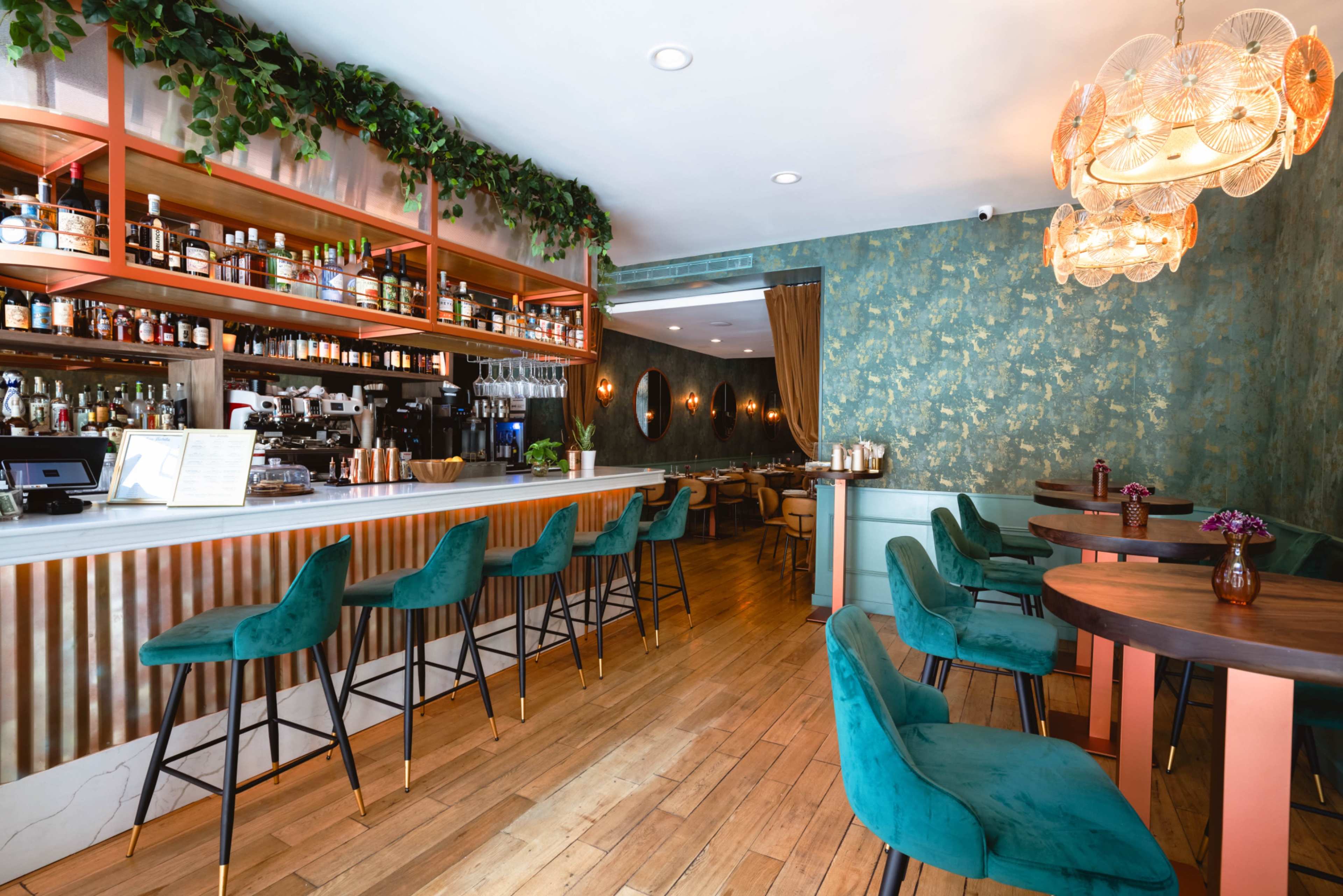 The image shows a modern bar area with stylish green upholstered chairs, a well-stocked shelf of liquor, and elegant lighting fixtures in a restaurant setting.