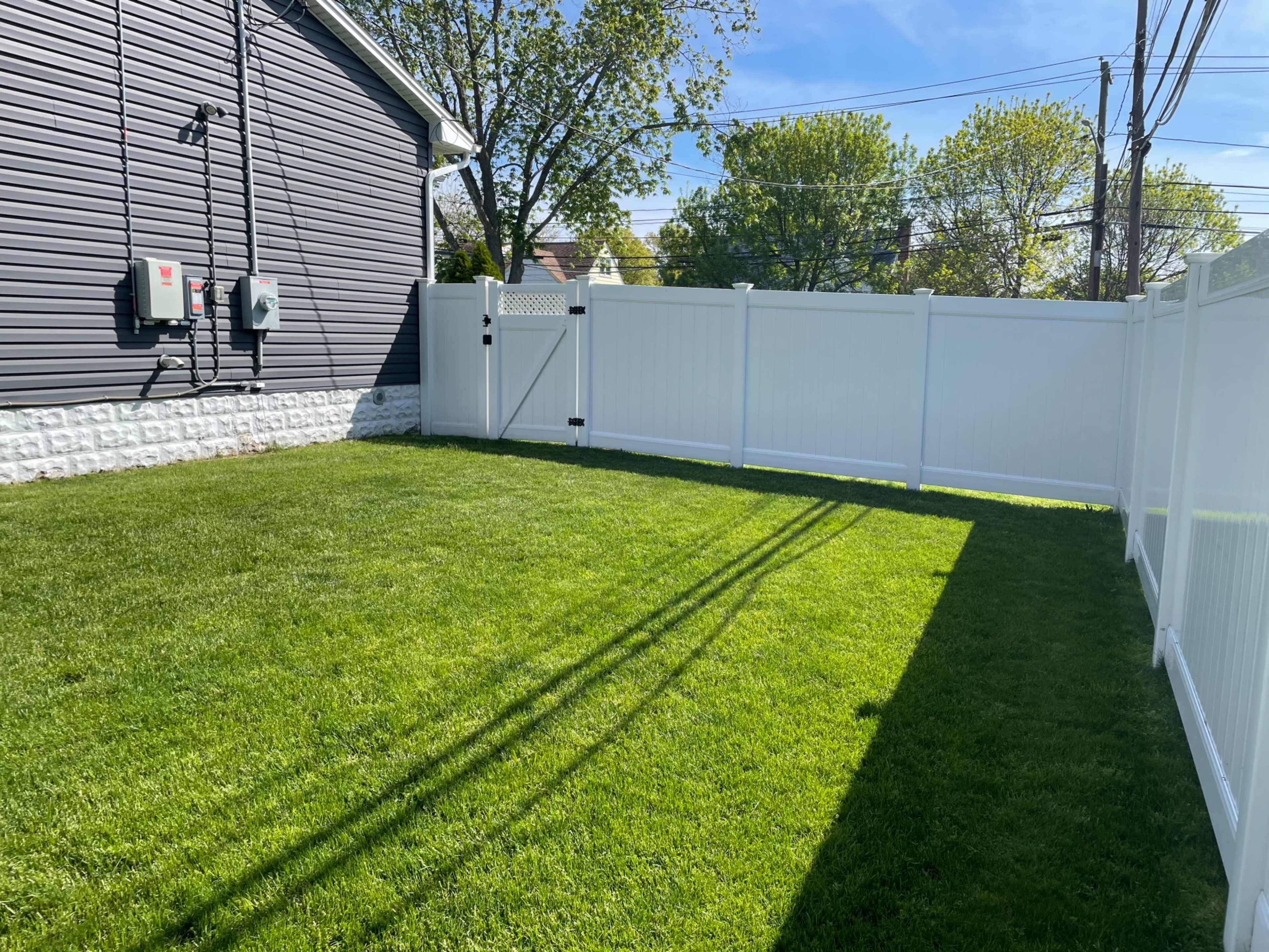 The image shows a neatly maintained grassy yard enclosed by a white vinyl fence, with shadow lines cast by nearby fence posts.