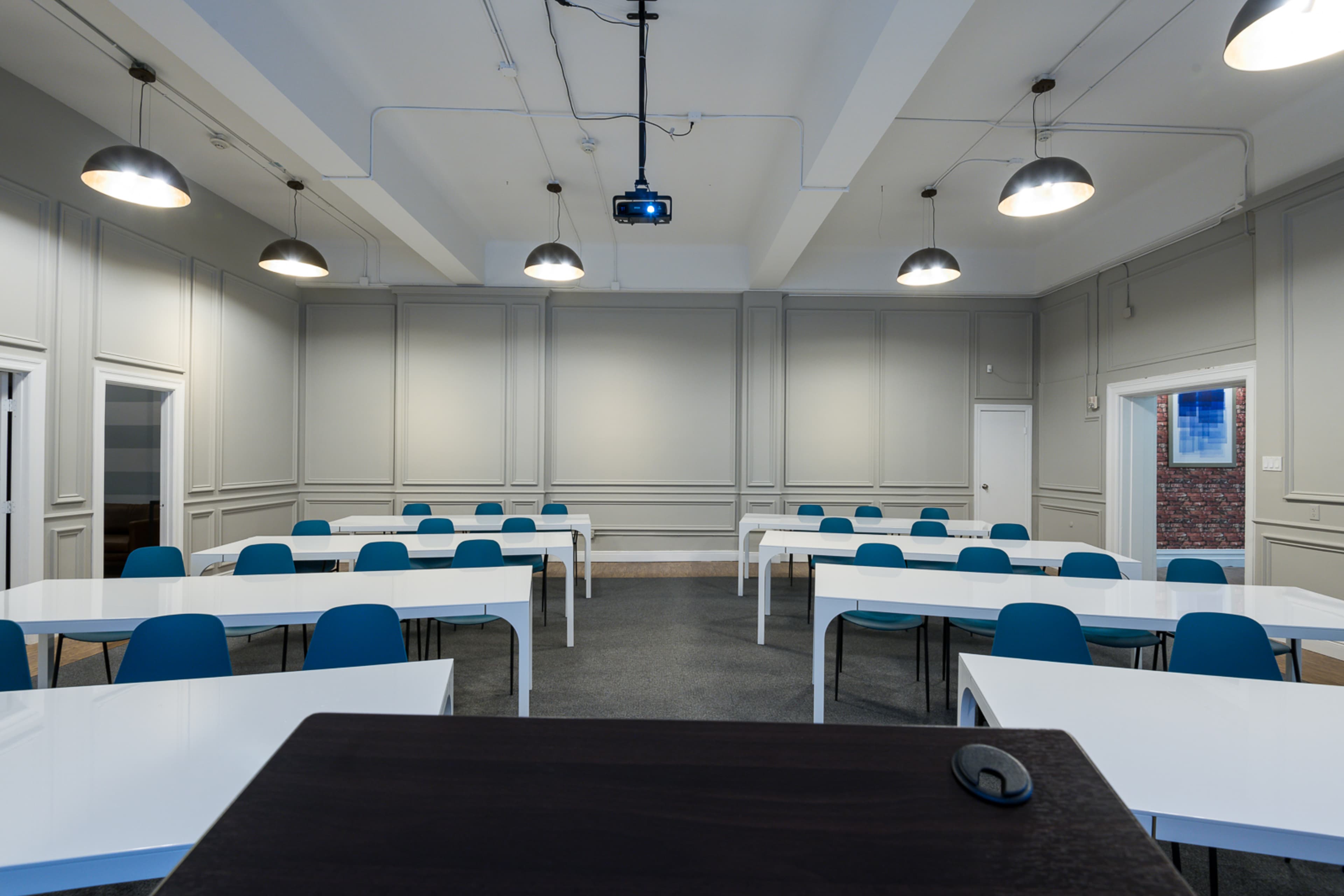 The image shows a classroom with several white tables arranged in rows and blue chairs, along with a projector hanging from the ceiling.