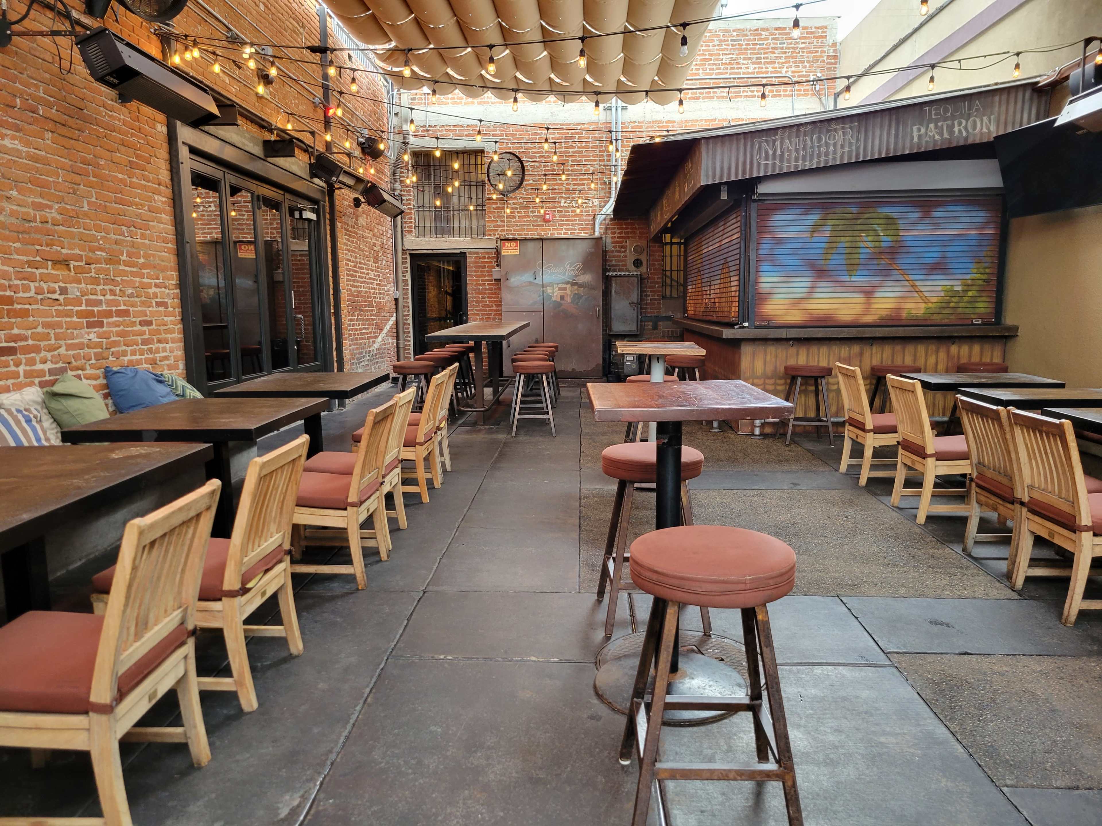 A patio area of a restaurant featuring wooden tables and chairs against a backdrop of brick walls and overhead string lights.