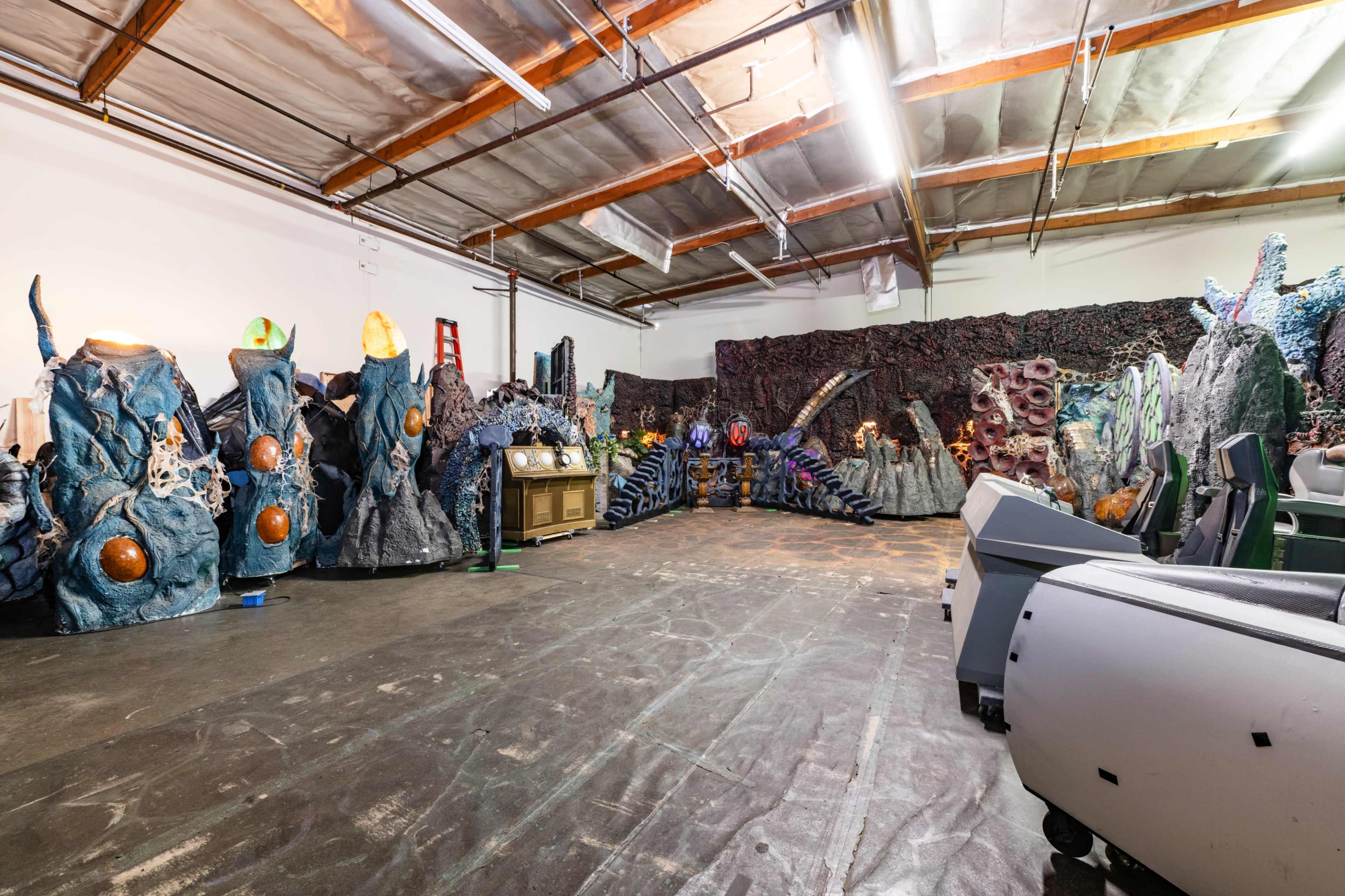 The image shows an industrial warehouse space filled with large, colorful stage props resembling sea creatures and coral formations.