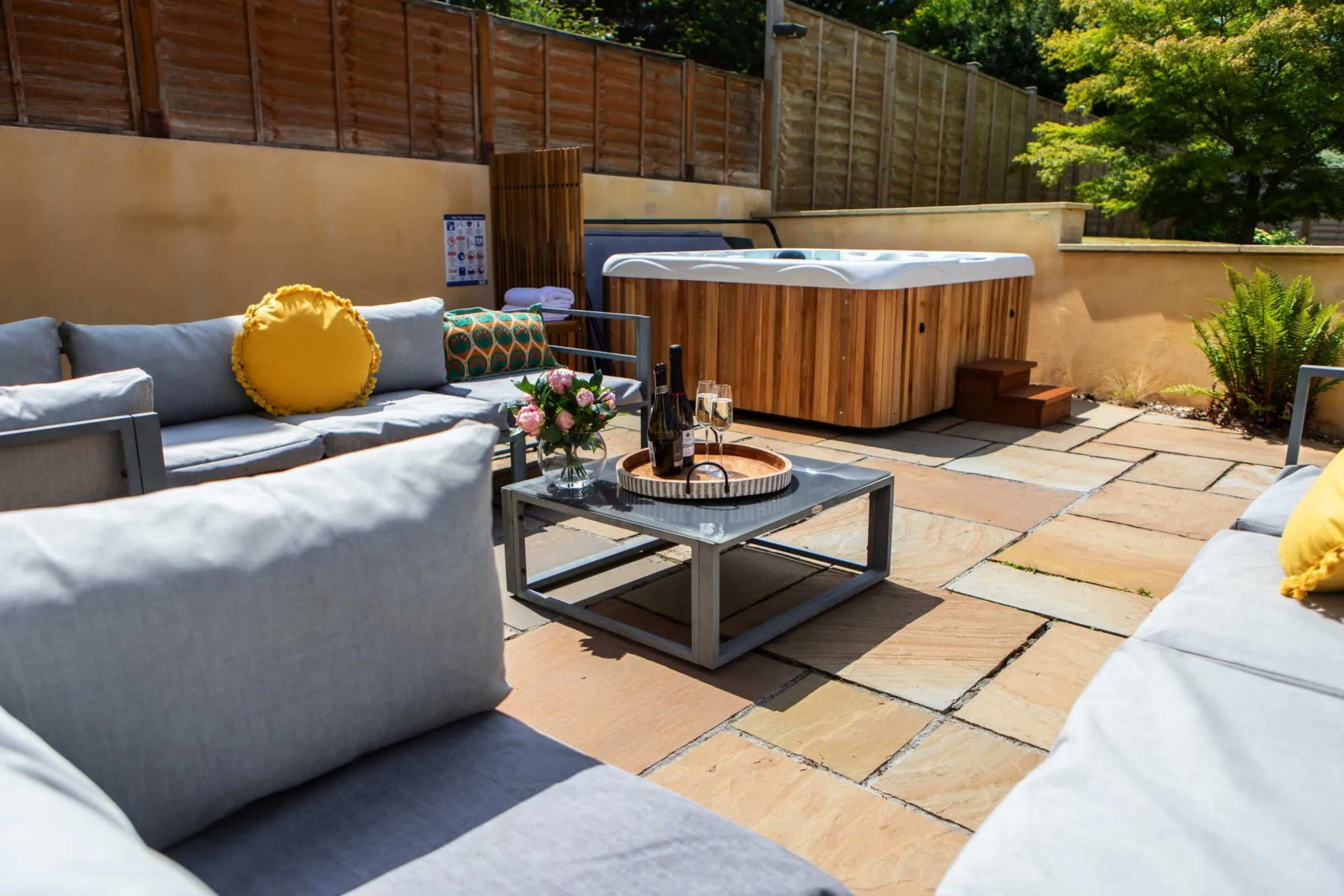 A patio area features a hot tub surrounded by outdoor seating, a coffee table with decorative items, and a backdrop of wood fencing and greenery.