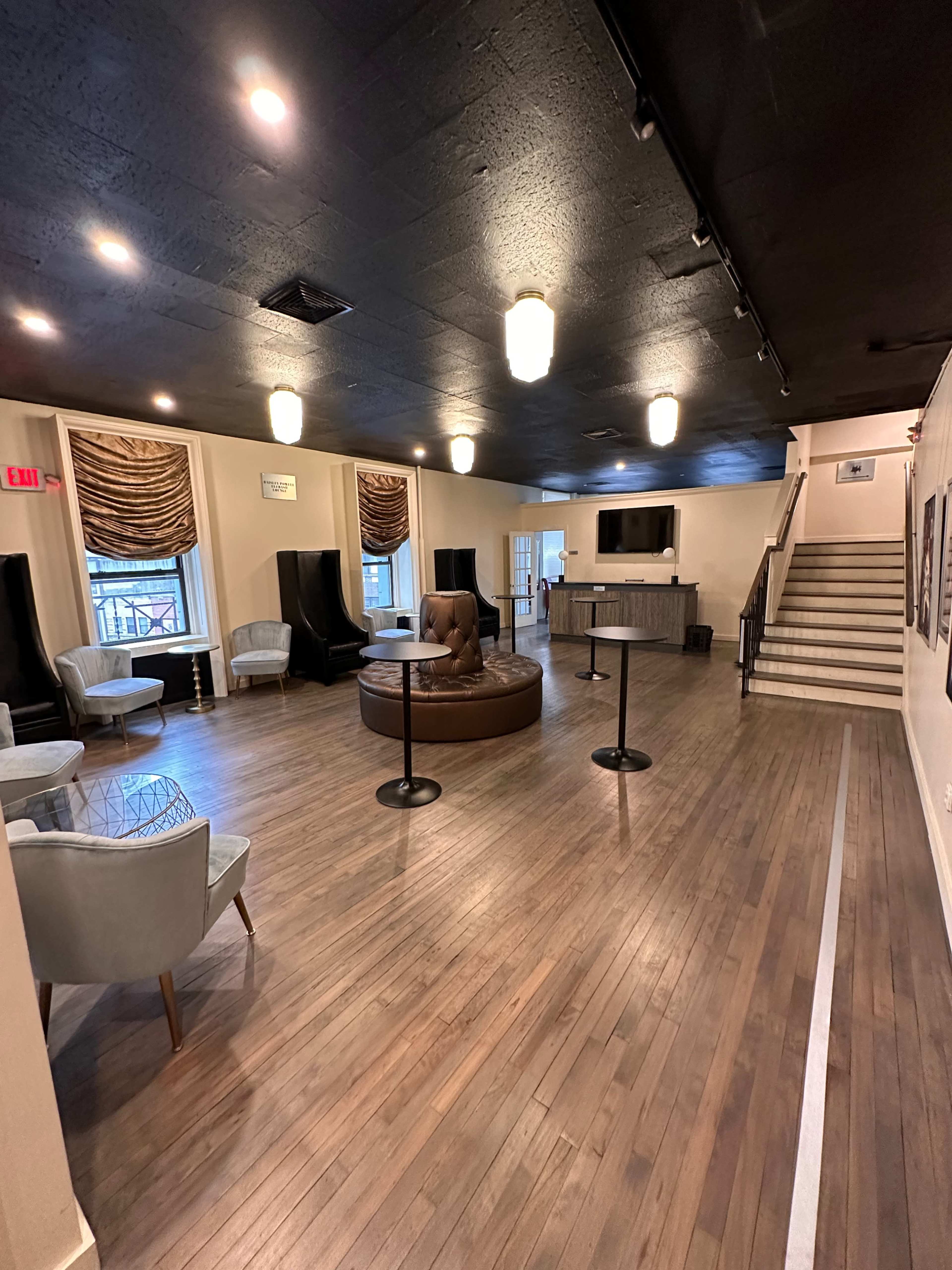 The image shows an empty lounge area with wooden floors, soft seating options, a large circular seating arrangement, and a staircase leading to an upper level.