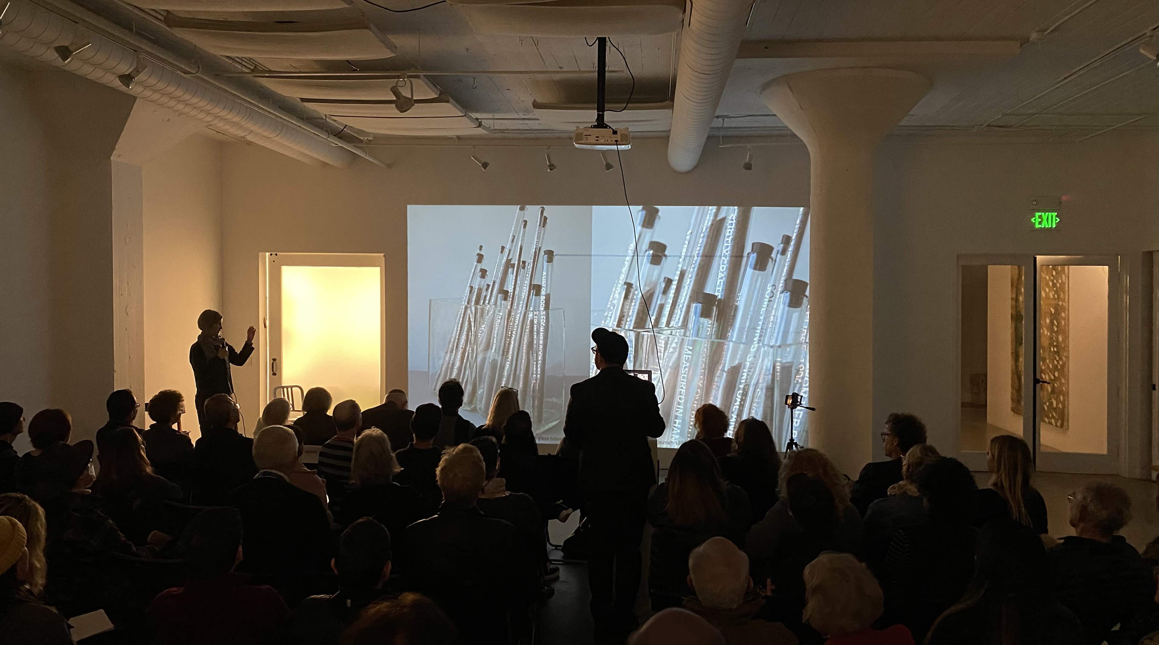 A speaker presents to an attentive audience as images of flutes are projected on a large screen in an art gallery setting.
