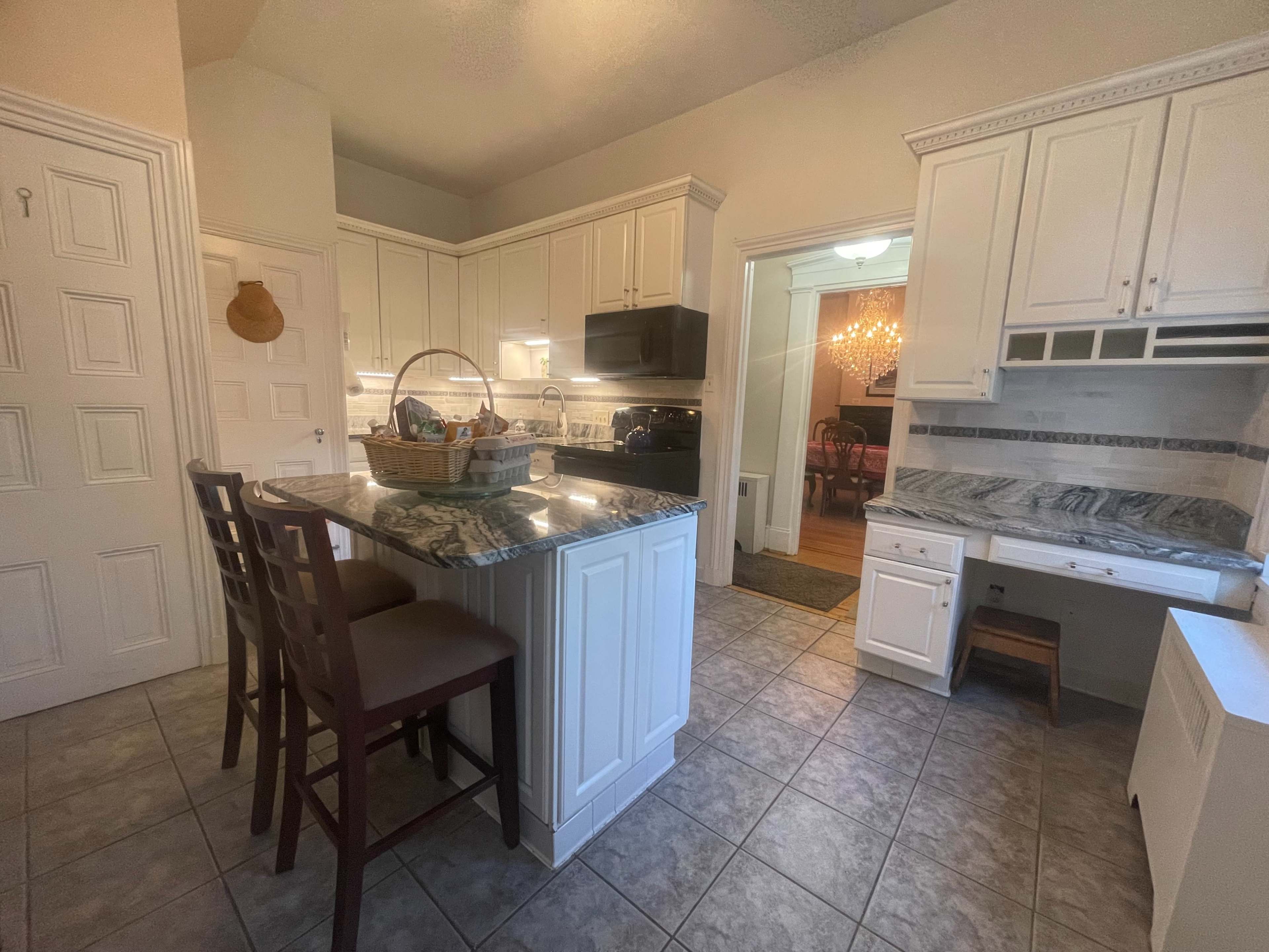 A well-lit kitchen features white cabinetry, a granite countertop island with seating, and tiled flooring.