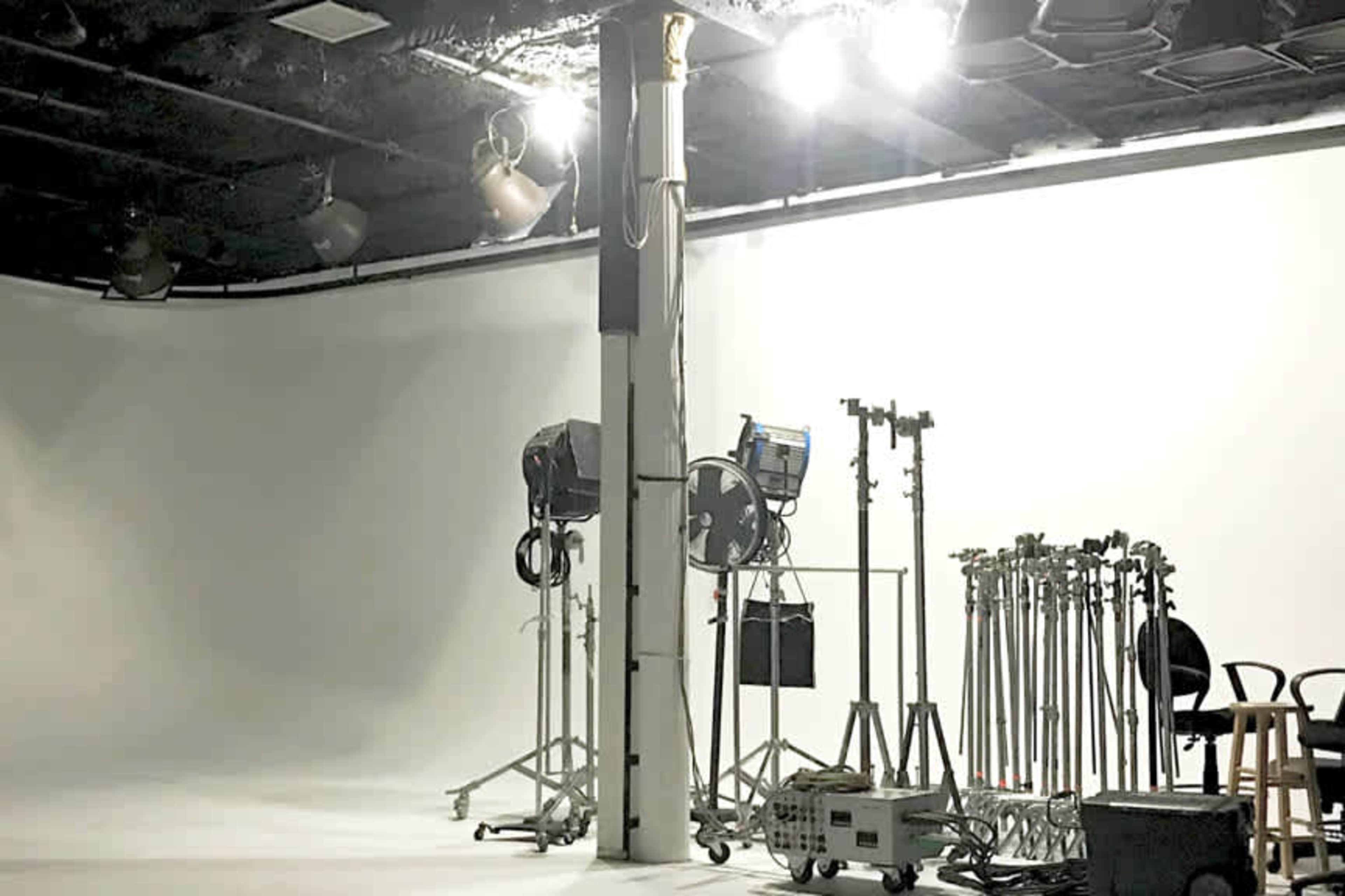 The image shows a photography or filming studio with a blank white backdrop, lighting equipment, and various stands arranged around a central column.