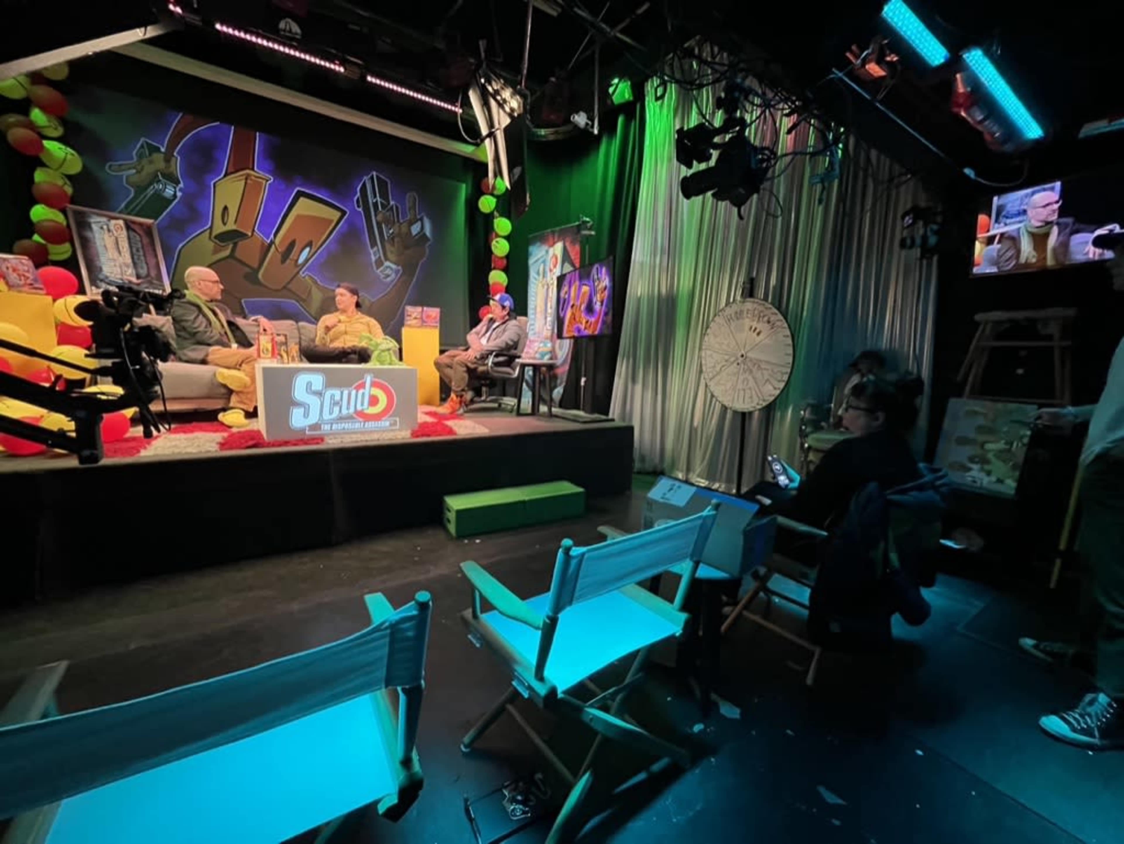 The image shows a television studio set with colorful backdrops and seating, where two guests are seated for a talk show, and crew members are visible in the foreground and background.