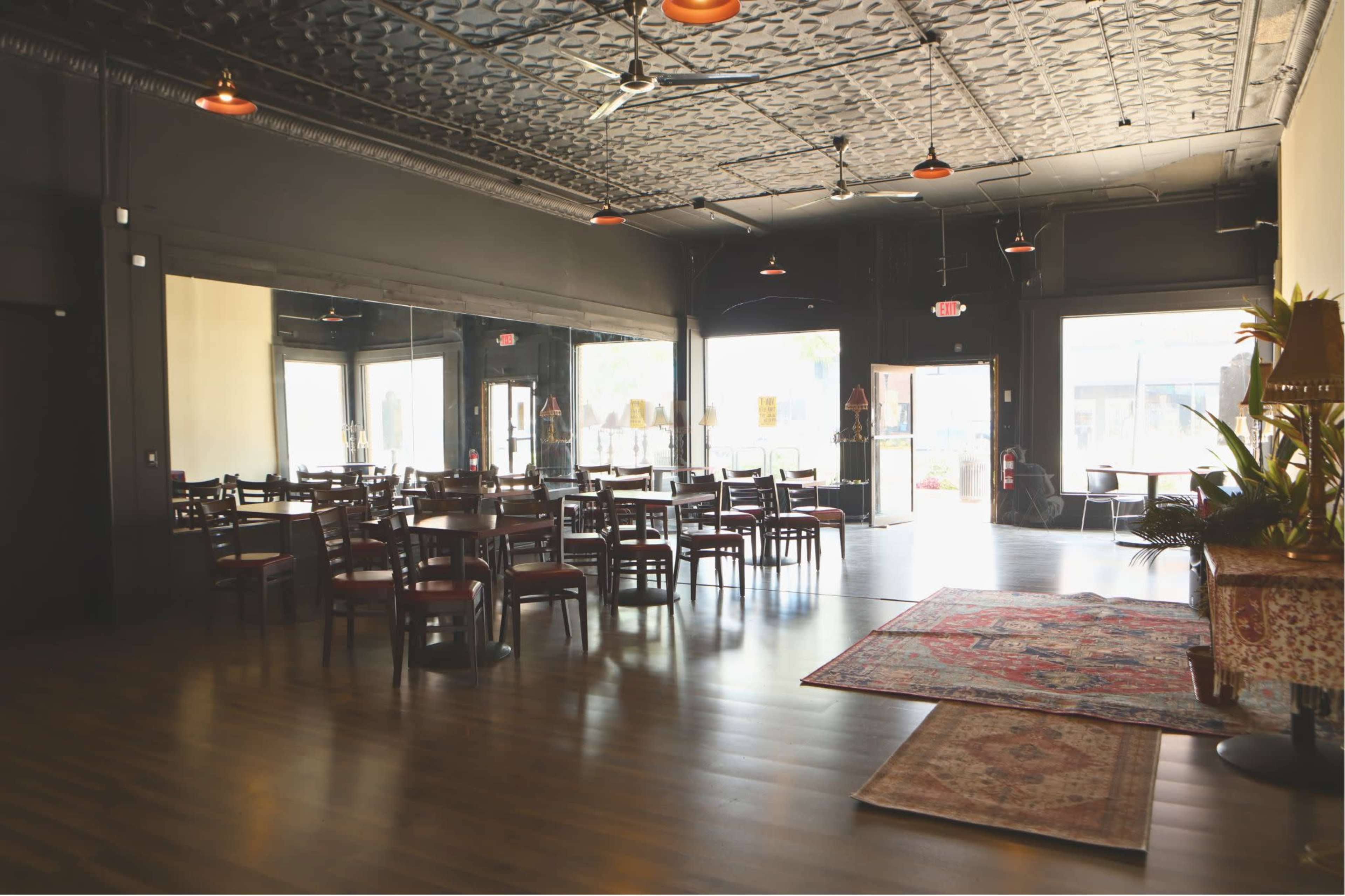 The image shows an empty restaurant interior with wooden tables and chairs, a patterned rug, and large windows letting in natural light.