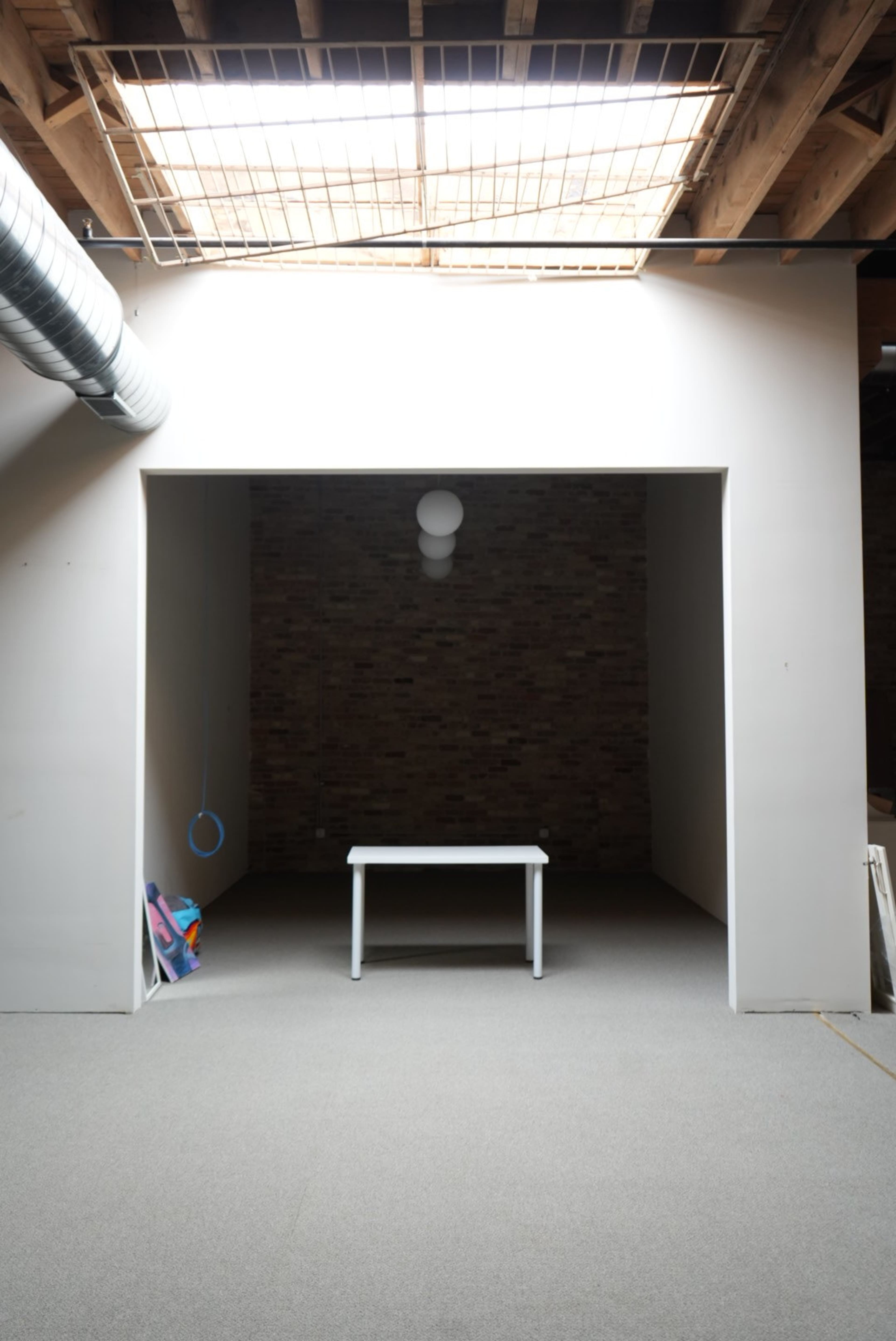 A minimalistic room features a small white table centered in an alcove, with brick walls and a skylight overhead.