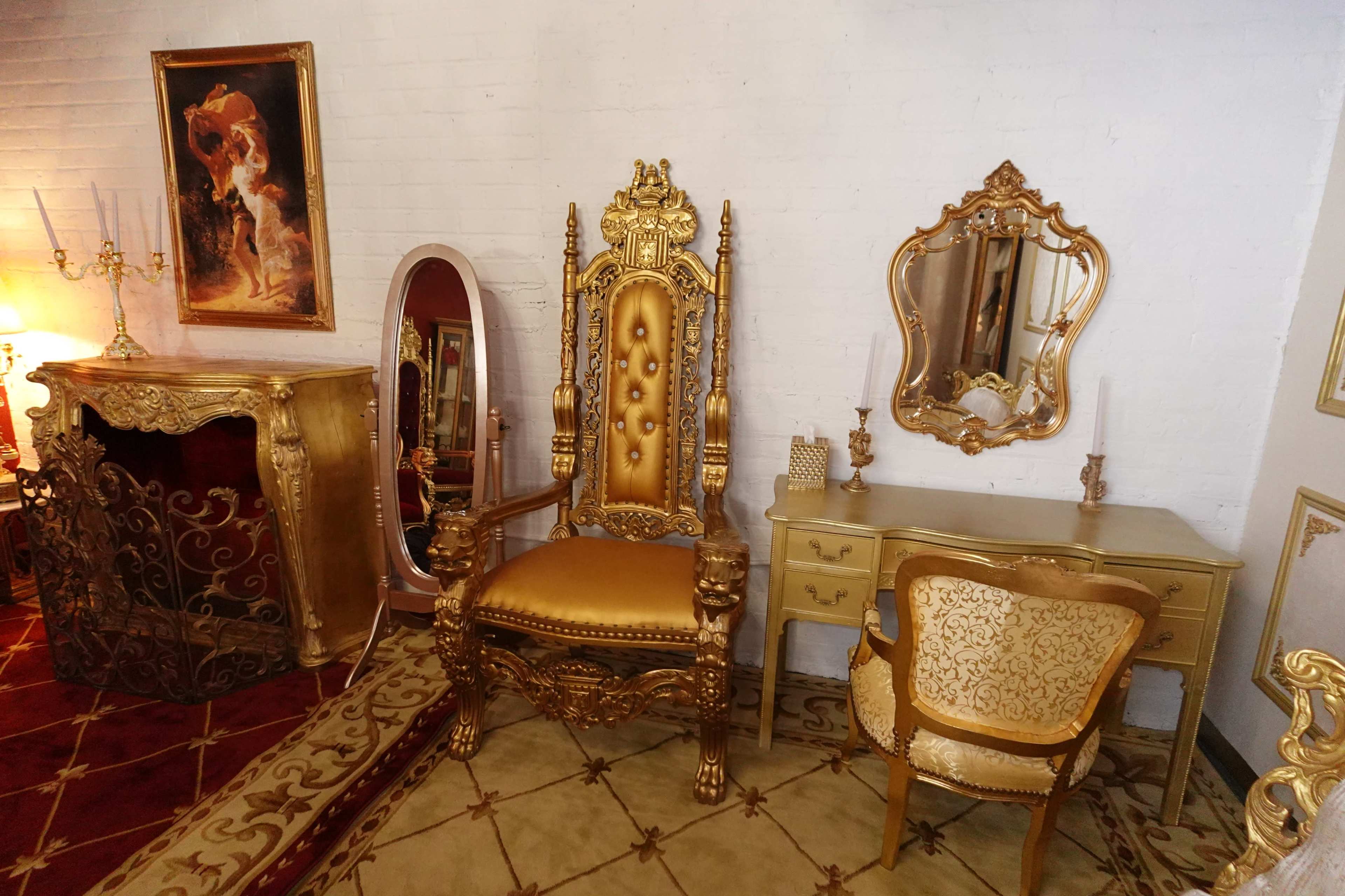The image shows an ornate room featuring a large, gold-accented throne chair, a gilded mirror, a decorative console table, and an intricately designed side chair, all set against a white brick wall.