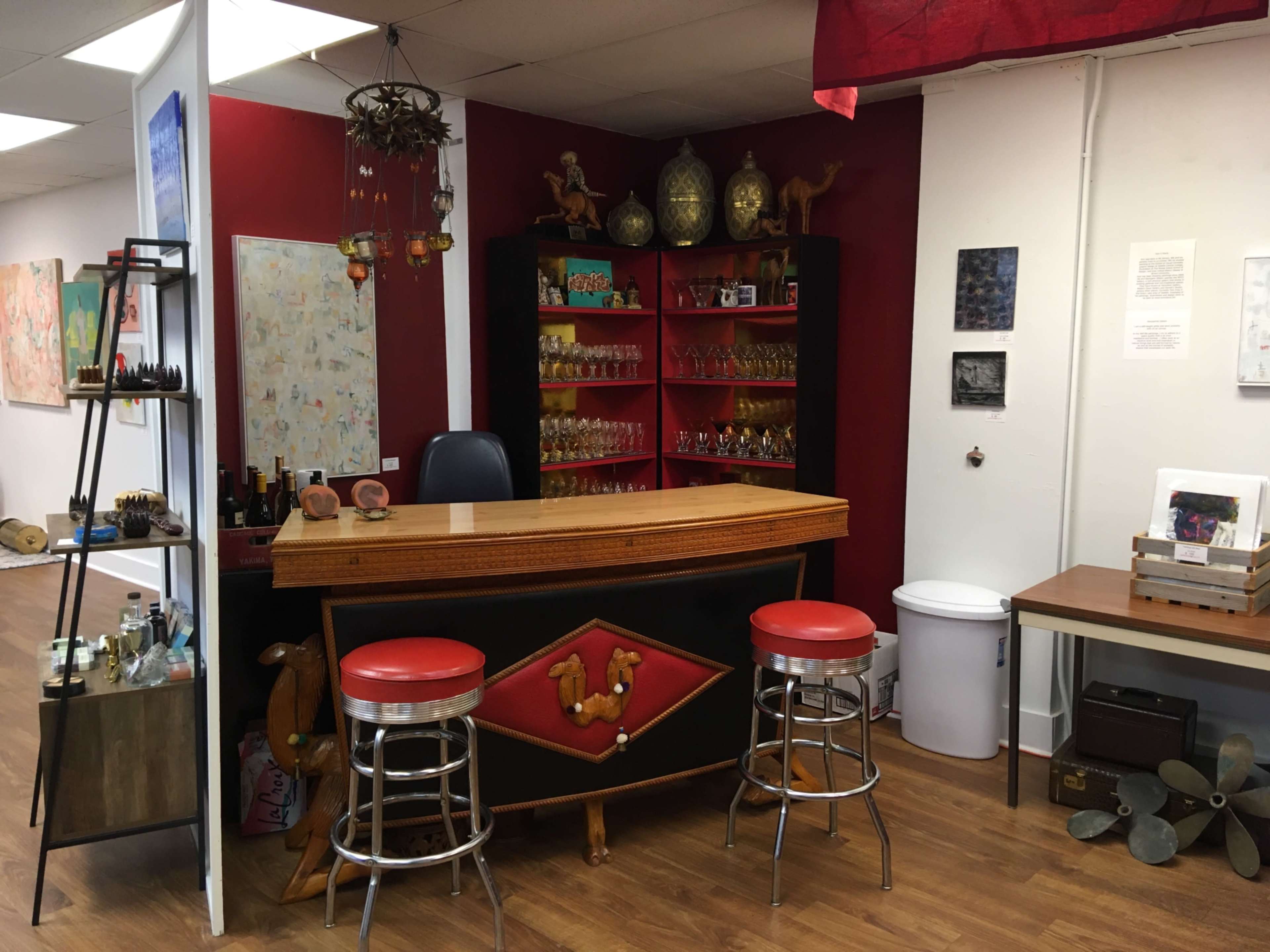 A bar area with a wooden counter, red stools, shelves filled with glassware, and decorative items against a backdrop of red and white walls.