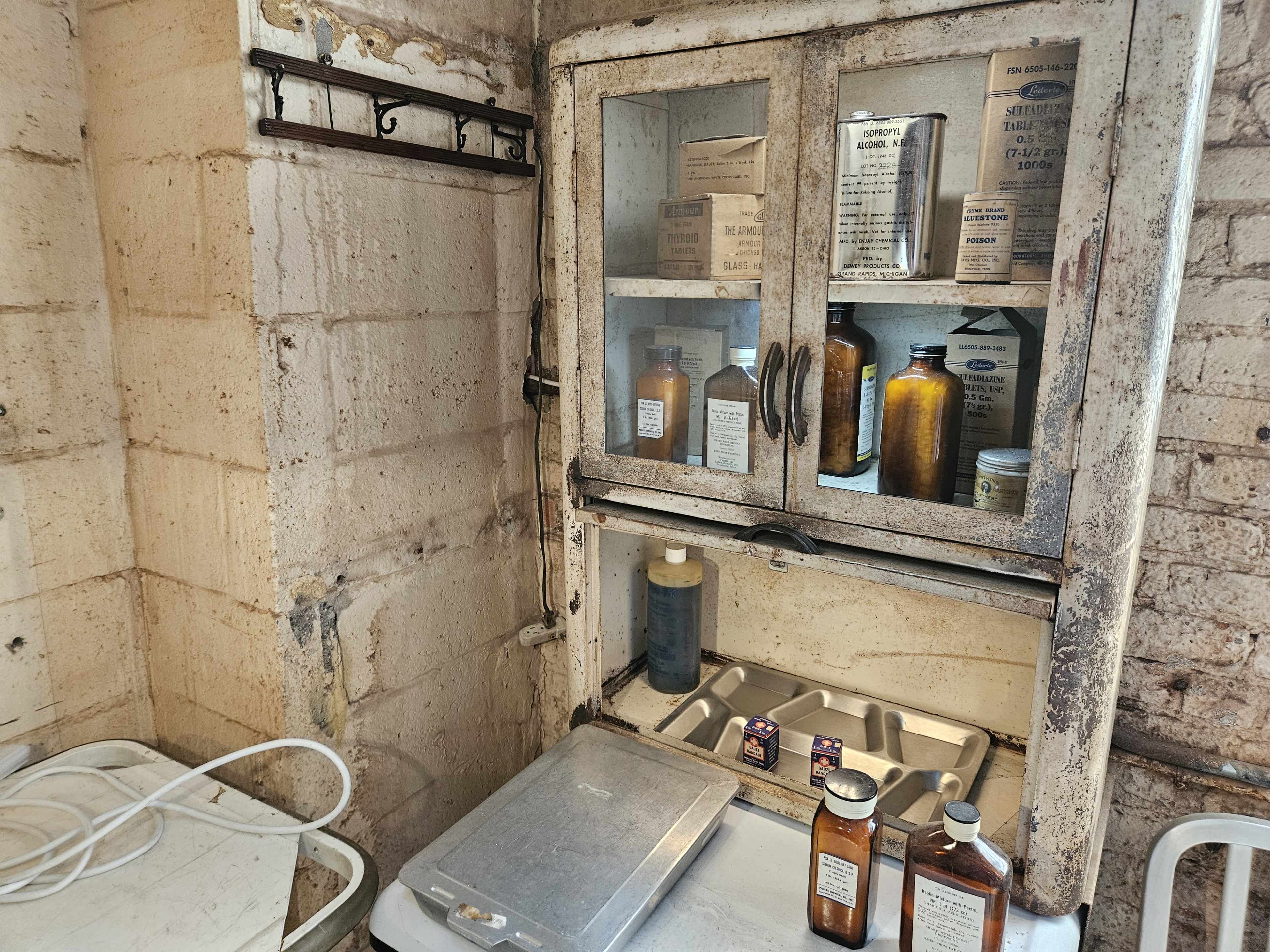 An old, weathered cabinet with glass doors displays various bottles and boxes, set against a bare brick wall in a dimly lit room.