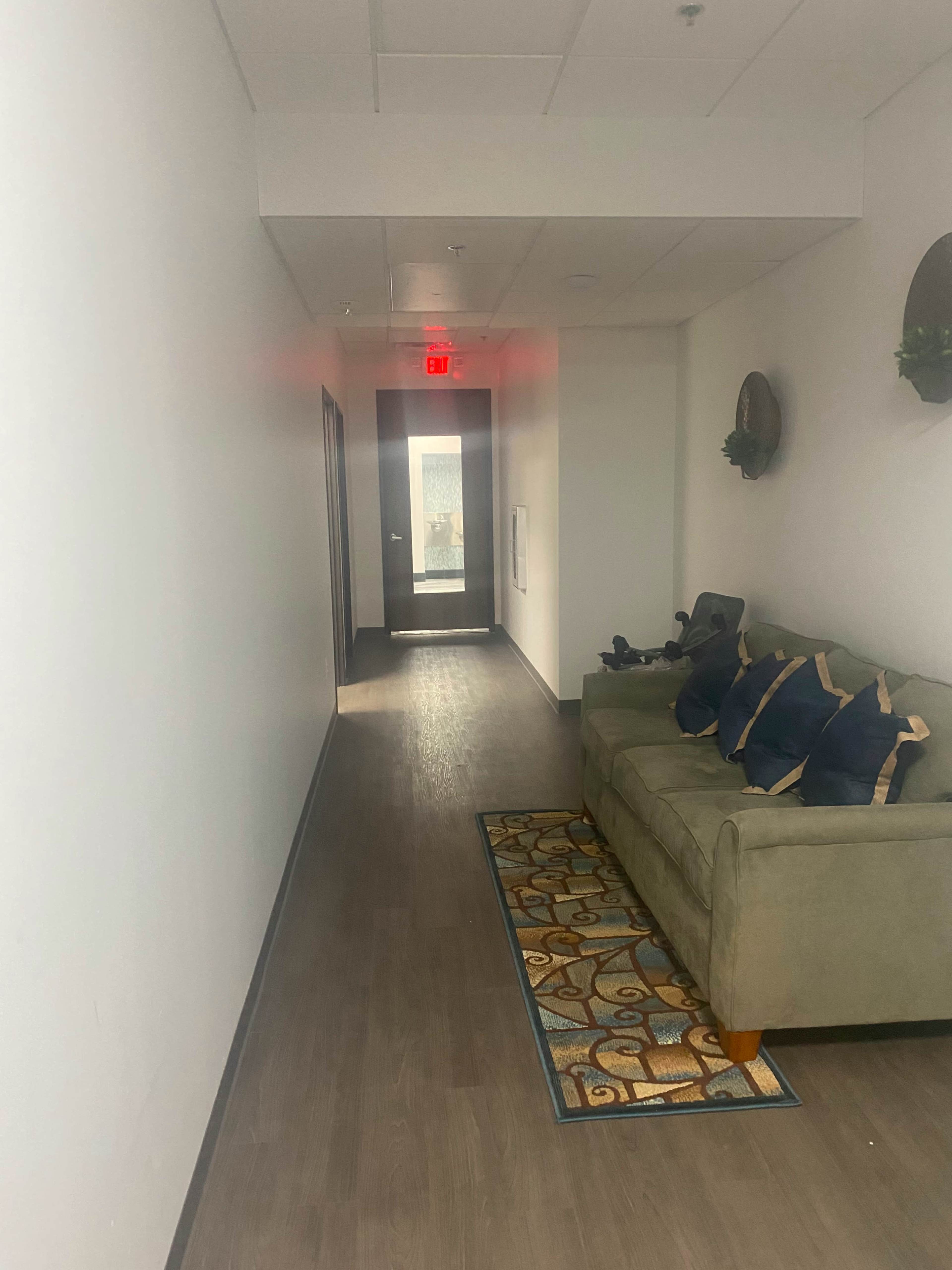 A narrow hallway with a green couch and decorative pillows on one side, leading to a glass door at the far end.