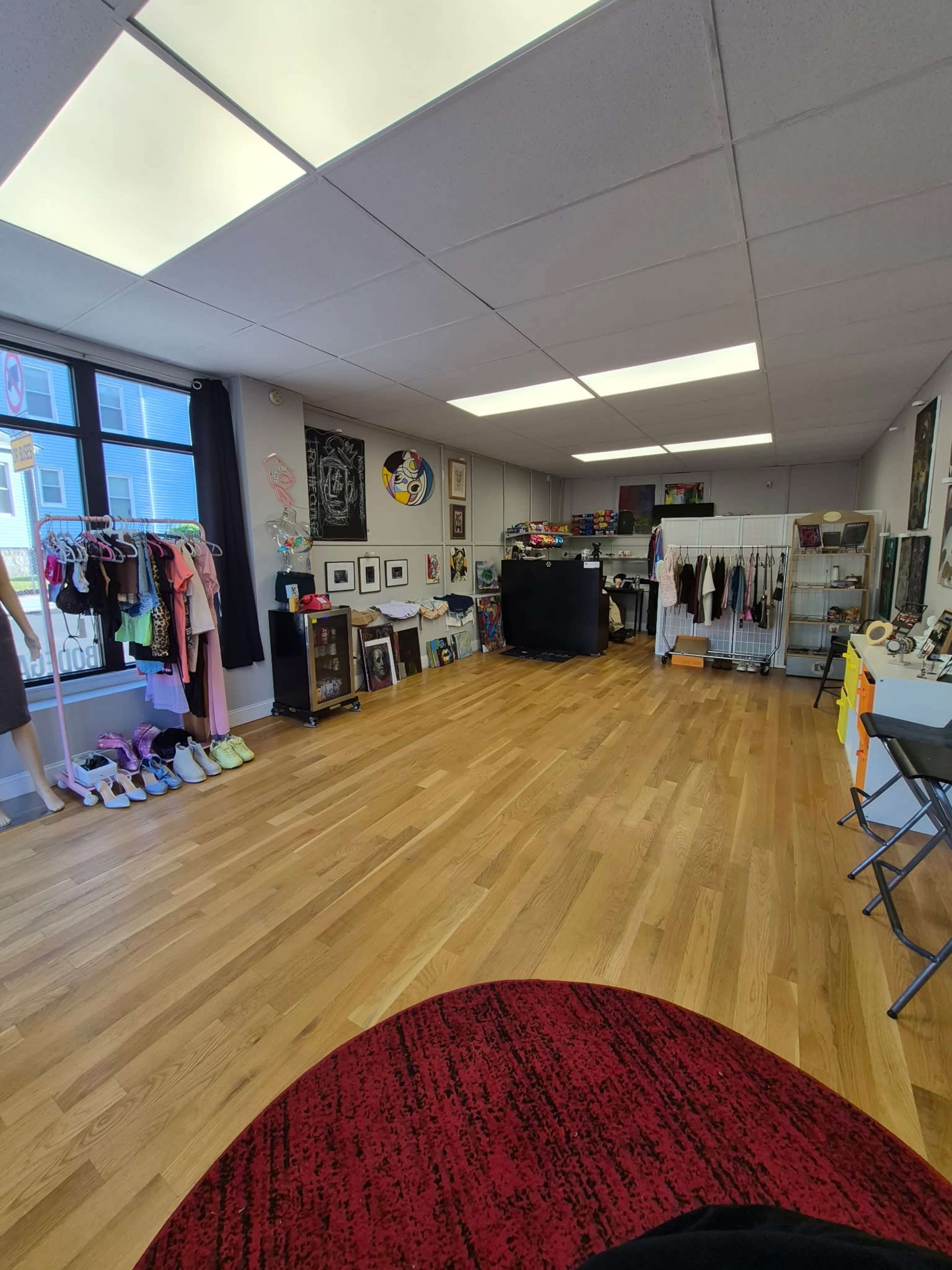 The image shows an interior of a retail space with wooden flooring, displaying clothing racks, art on the walls, and various items arranged in a clean, organized manner.