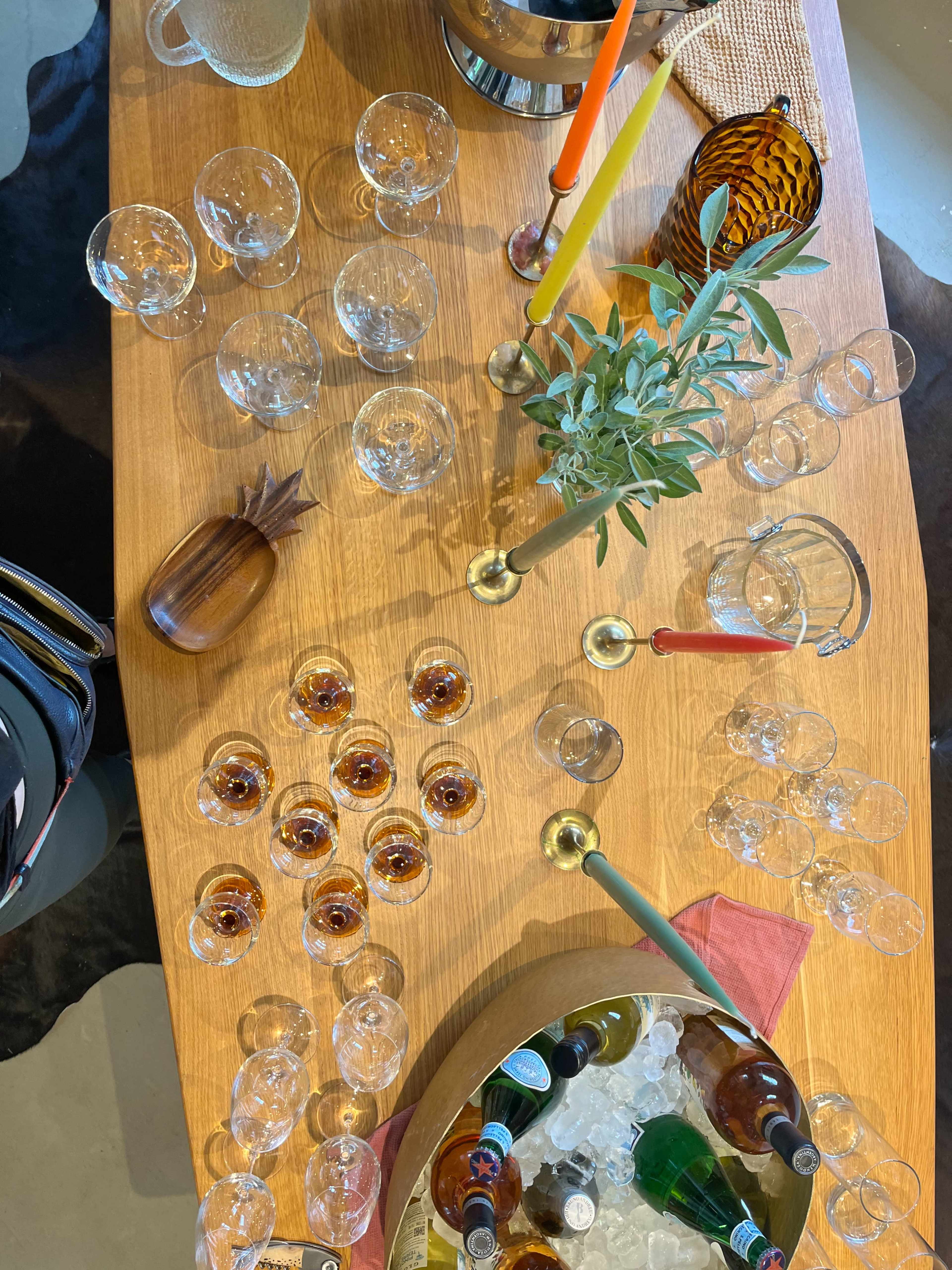 The image shows a wooden table set with various glassware, colorful candles, a bowl of ice with drinks, and a few decorative items.