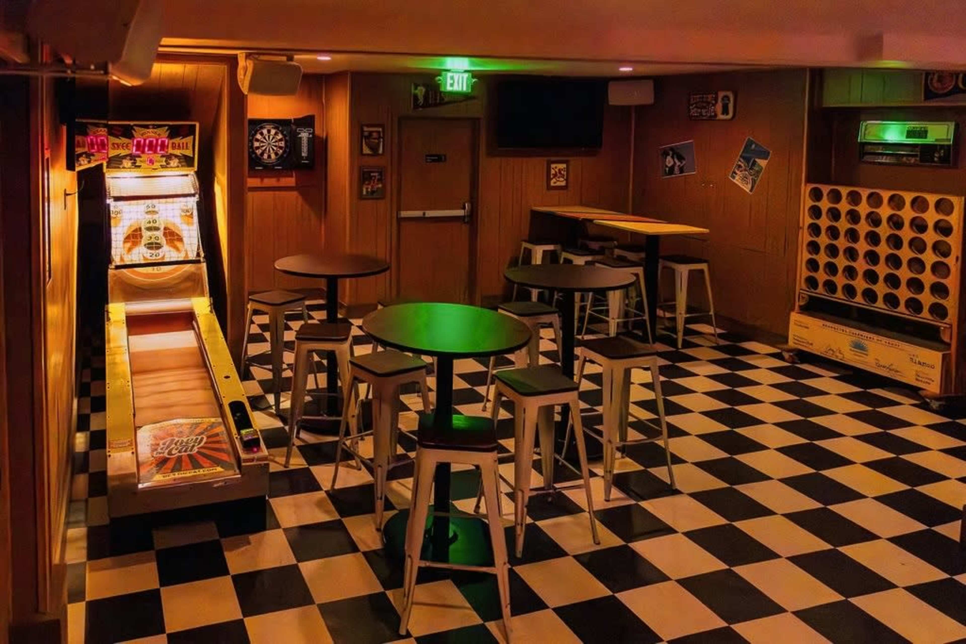 The image shows a dimly lit game room featuring a skee-ball machine, several small tables, and a checkered black-and-white floor.