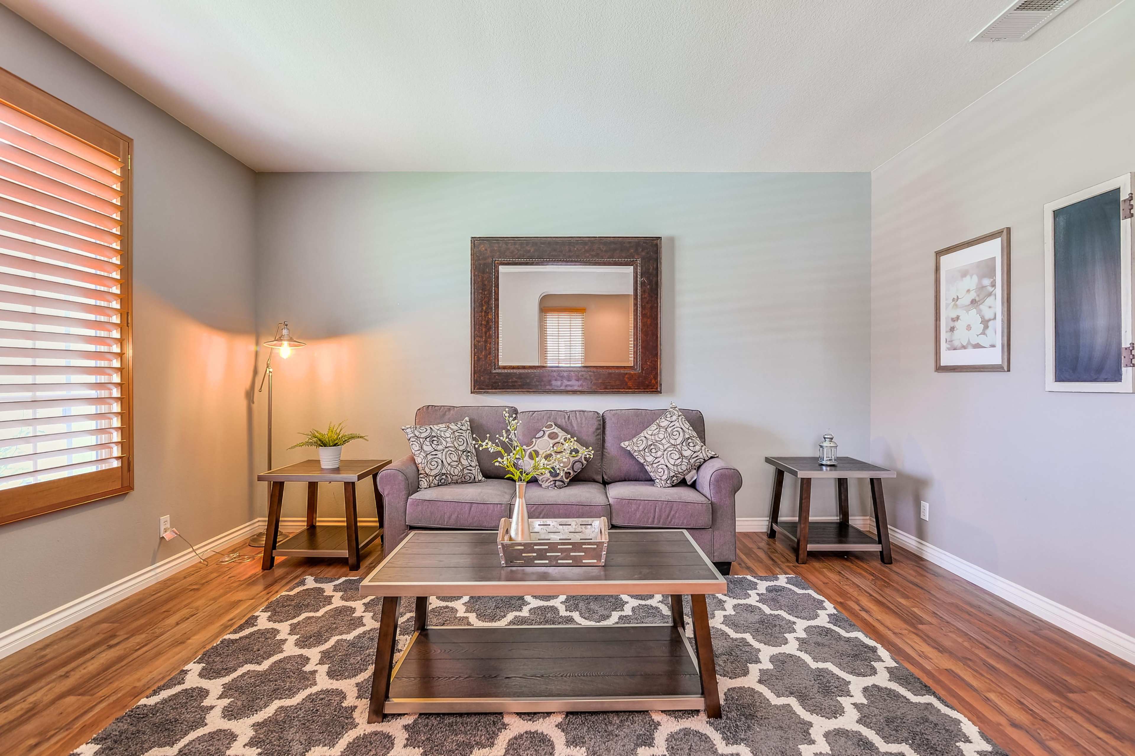 A living room with a purple sofa, two side tables, and a coffee table arranged on a patterned rug, illuminated by natural light through window blinds.