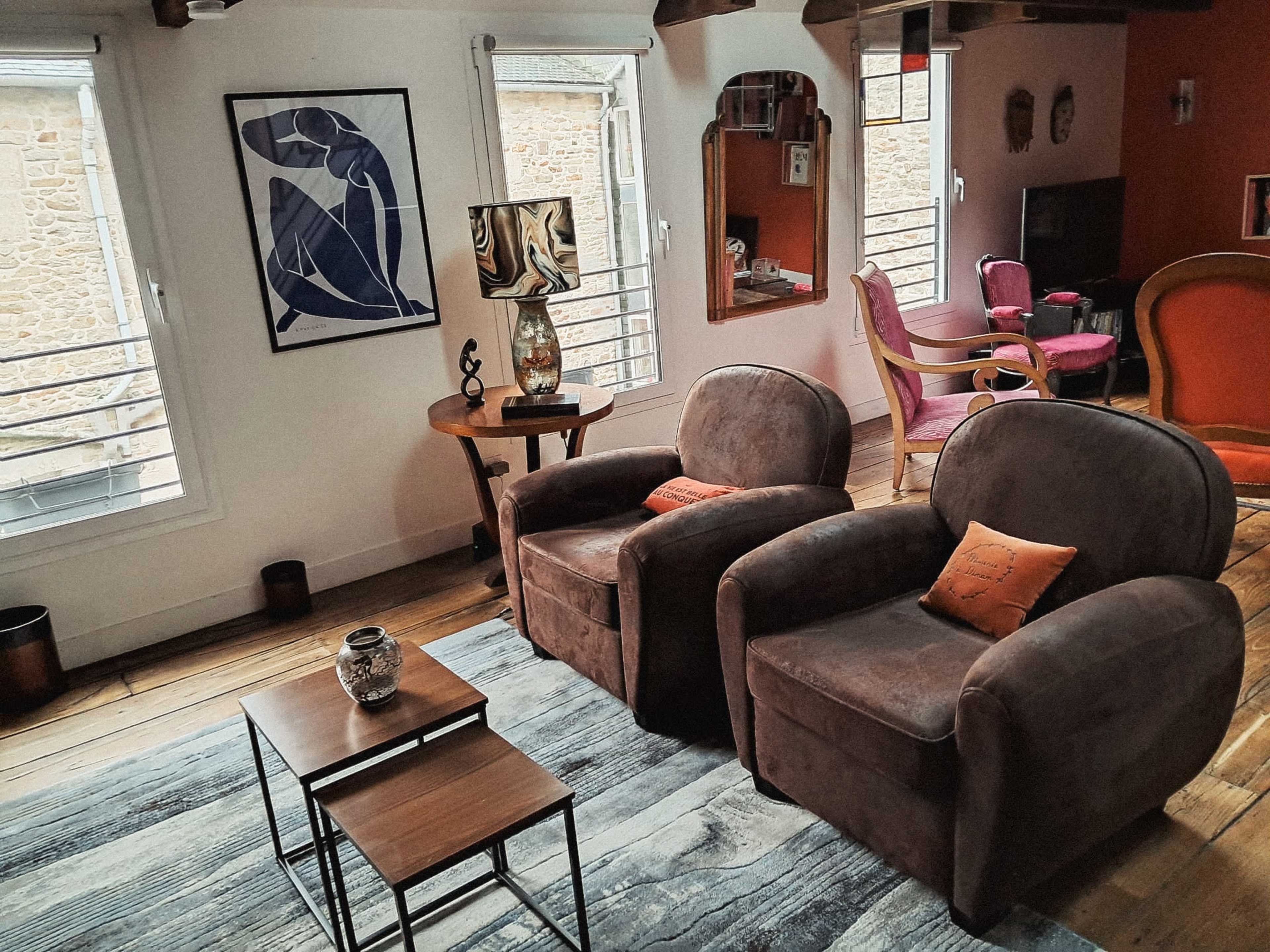 The scene features a cozy living room with two brown armchairs, a wooden coffee table, and decorative elements including a lamp and artwork on the walls.