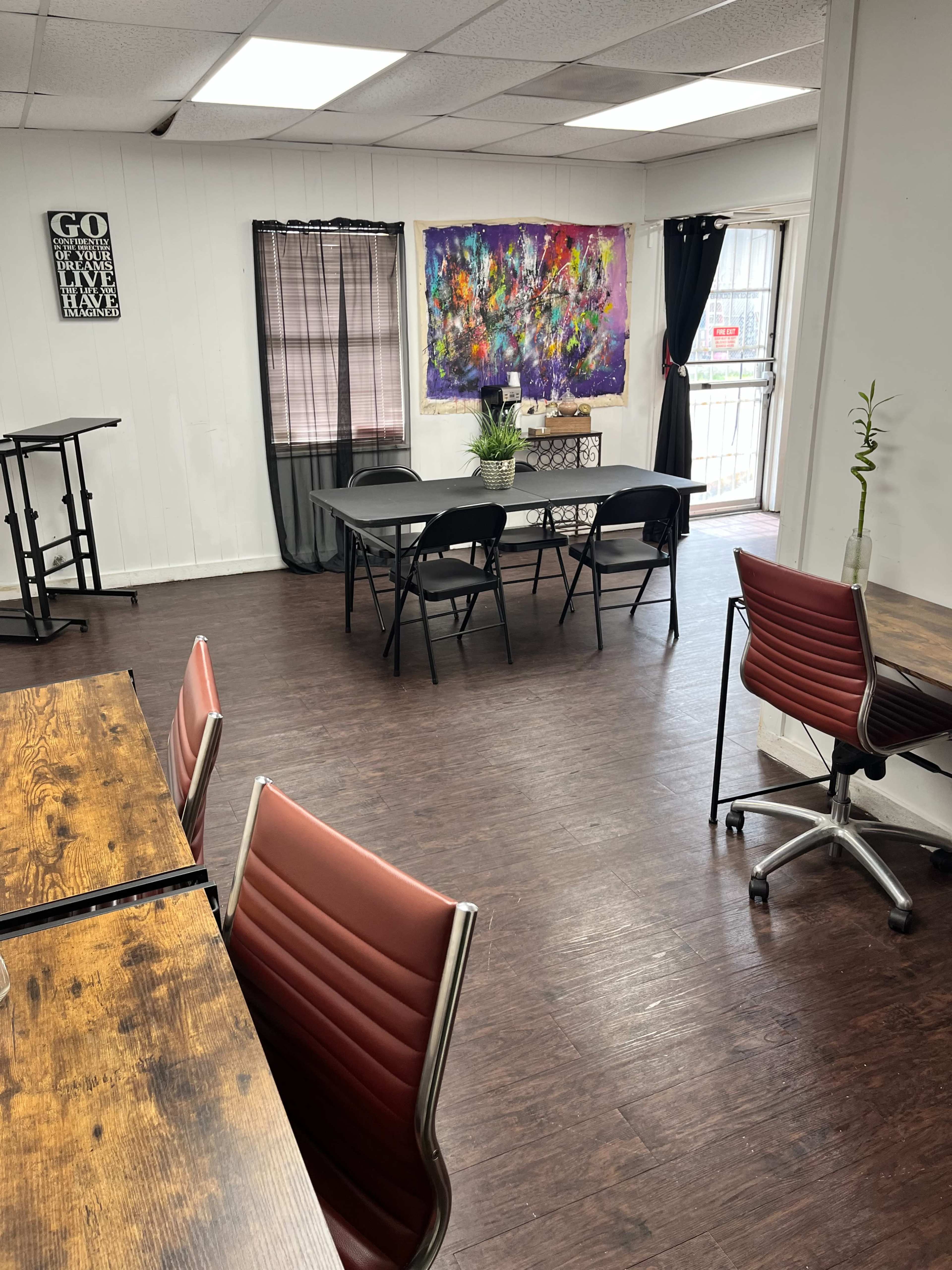 A spacious room with wooden flooring, featuring several tables and chairs, including a large table in the center and a colorful abstract painting on the wall.