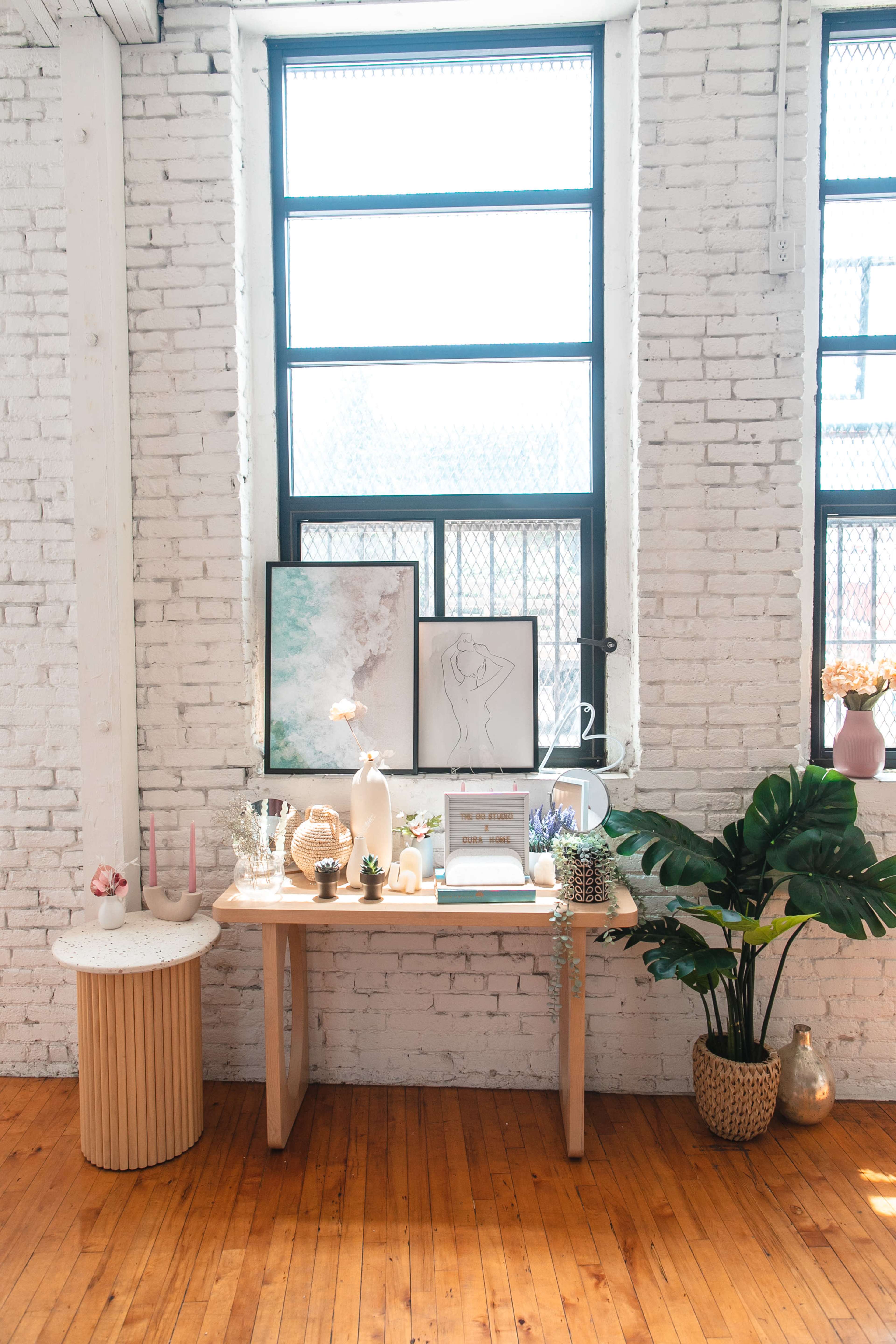 A wooden table with decorative items and plants sits against a wall of large windows in a bright, airy room.
