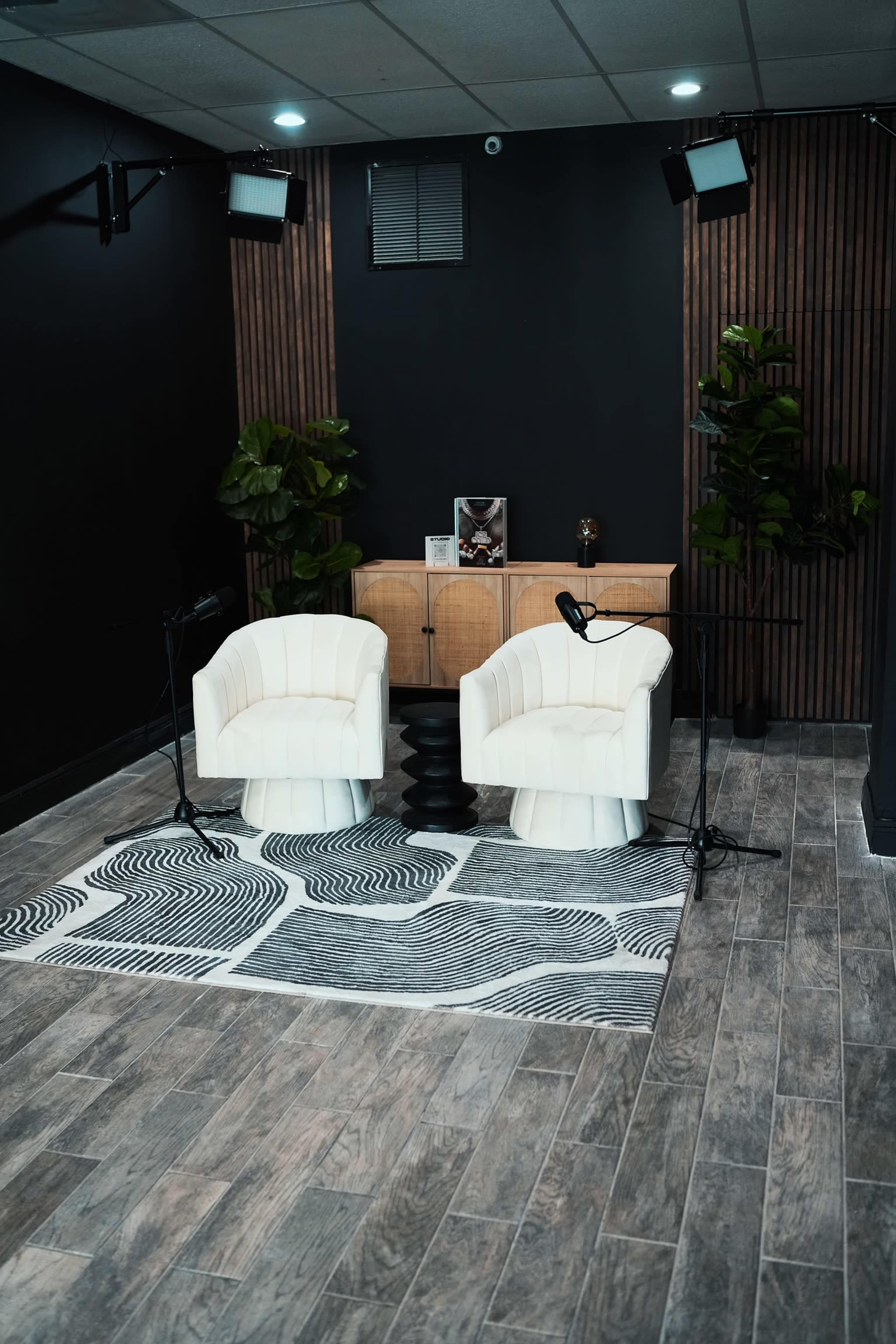 A modern studio space with two white chairs, a patterned rug, a wooden backdrop, and potted plants, accompanied by two microphones on stands.