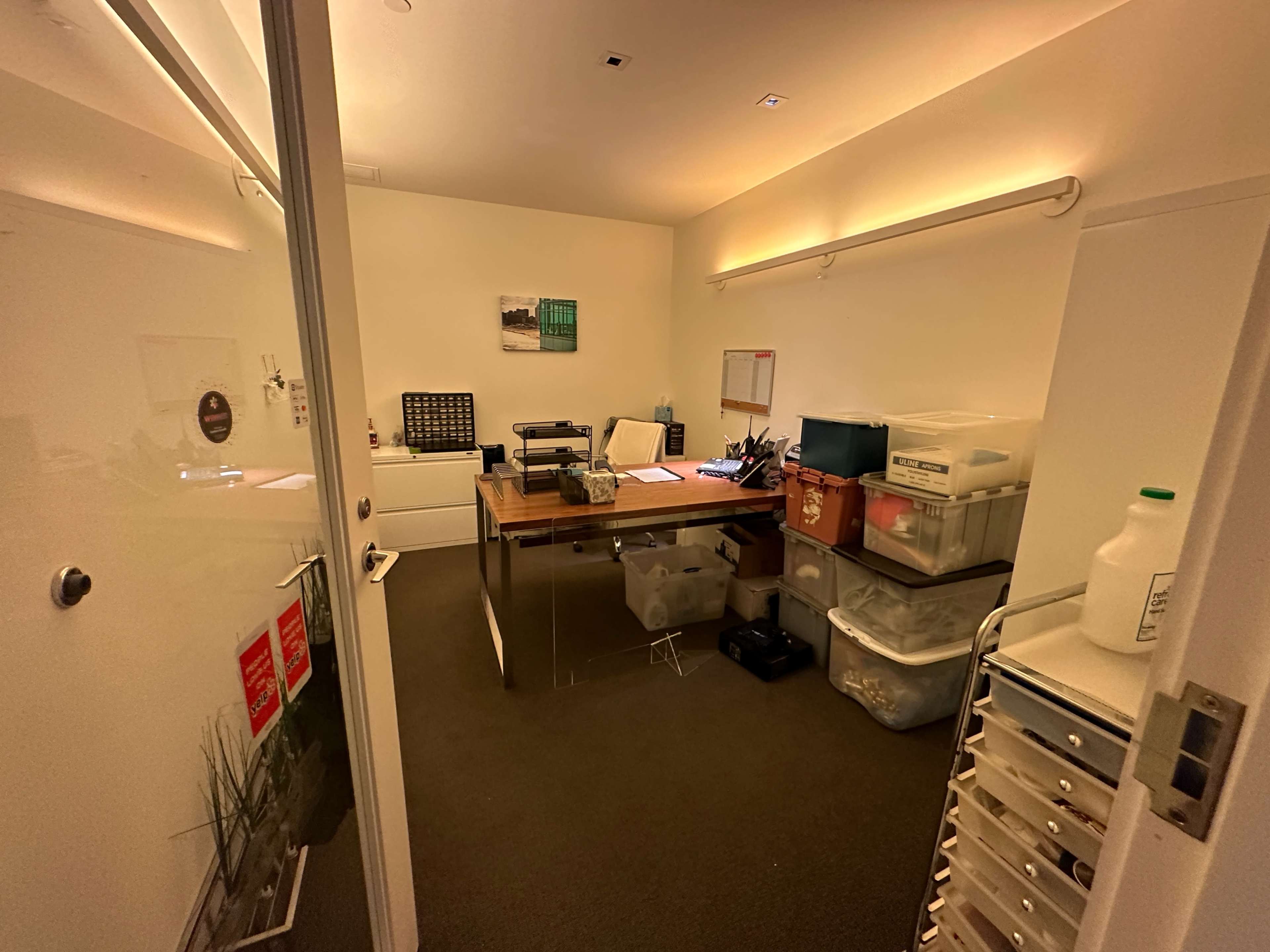 A small office space featuring a wooden table with office supplies, storage boxes, and a door leading to the corridor.