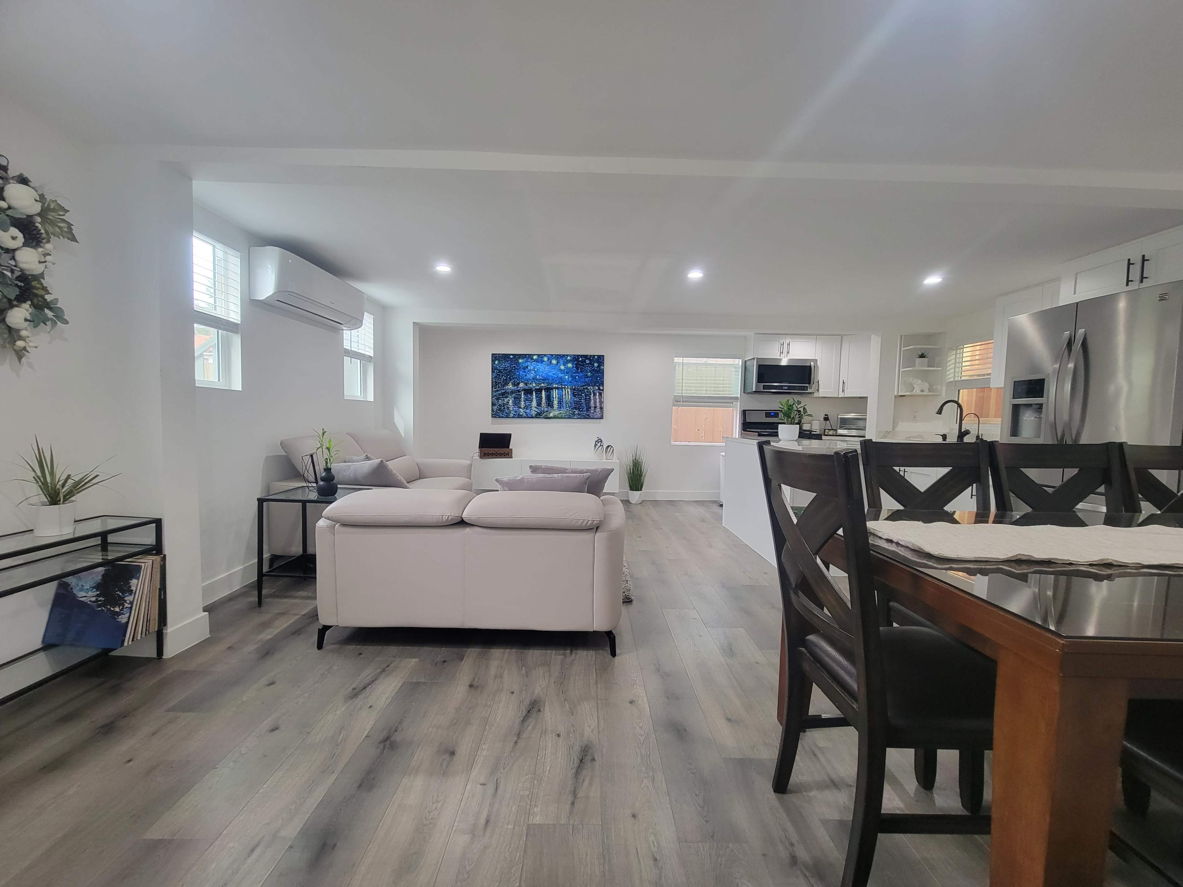 A modern, open-concept living space featuring a light-colored sofa, a dining area with a wooden table, and a kitchen in the background, all illuminated by natural light.