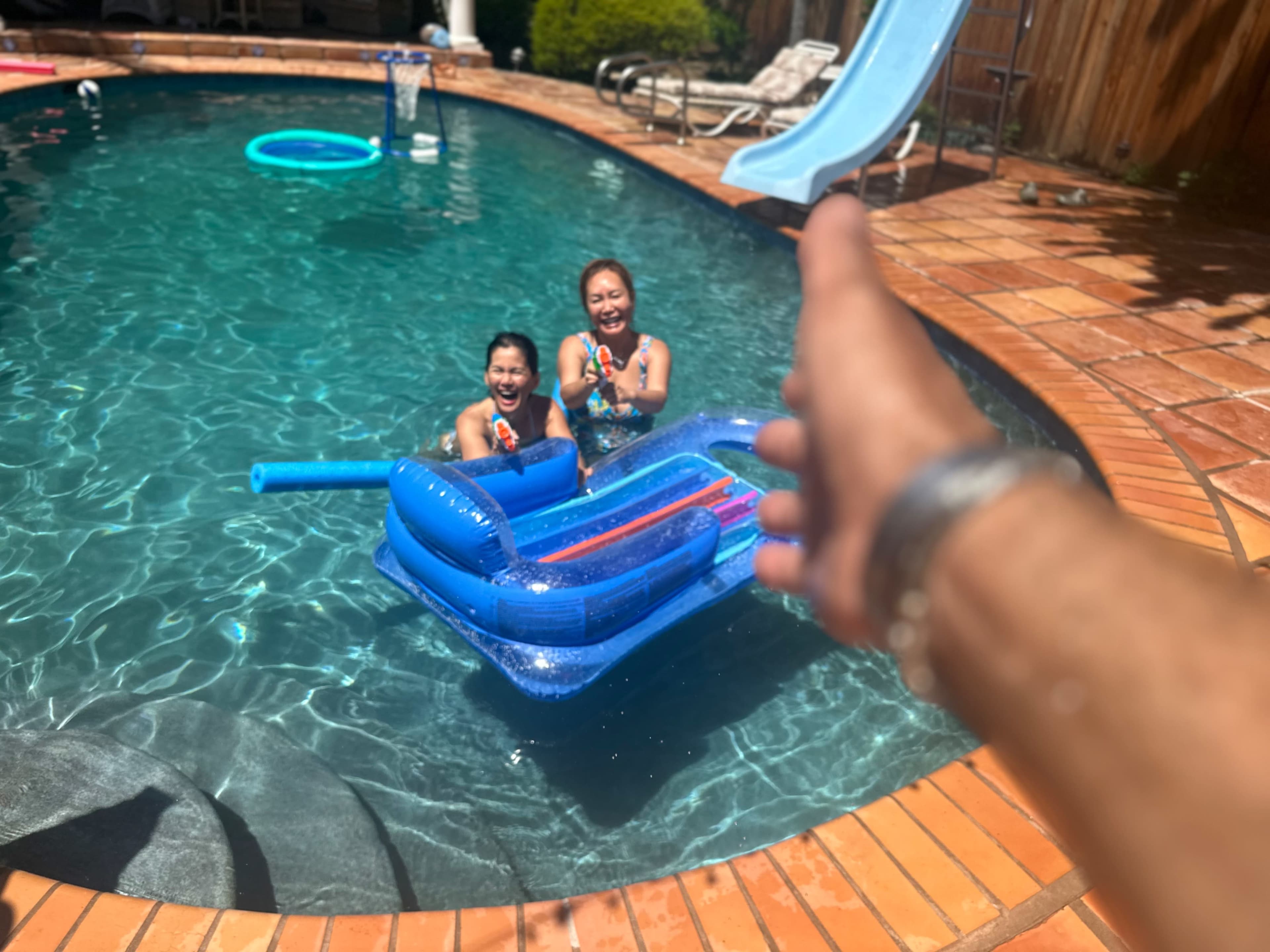 Two children sit on a blue inflatable float in a swimming pool, smiling and holding water guns, while an outstretched hand reaches toward them from the edge of the pool.