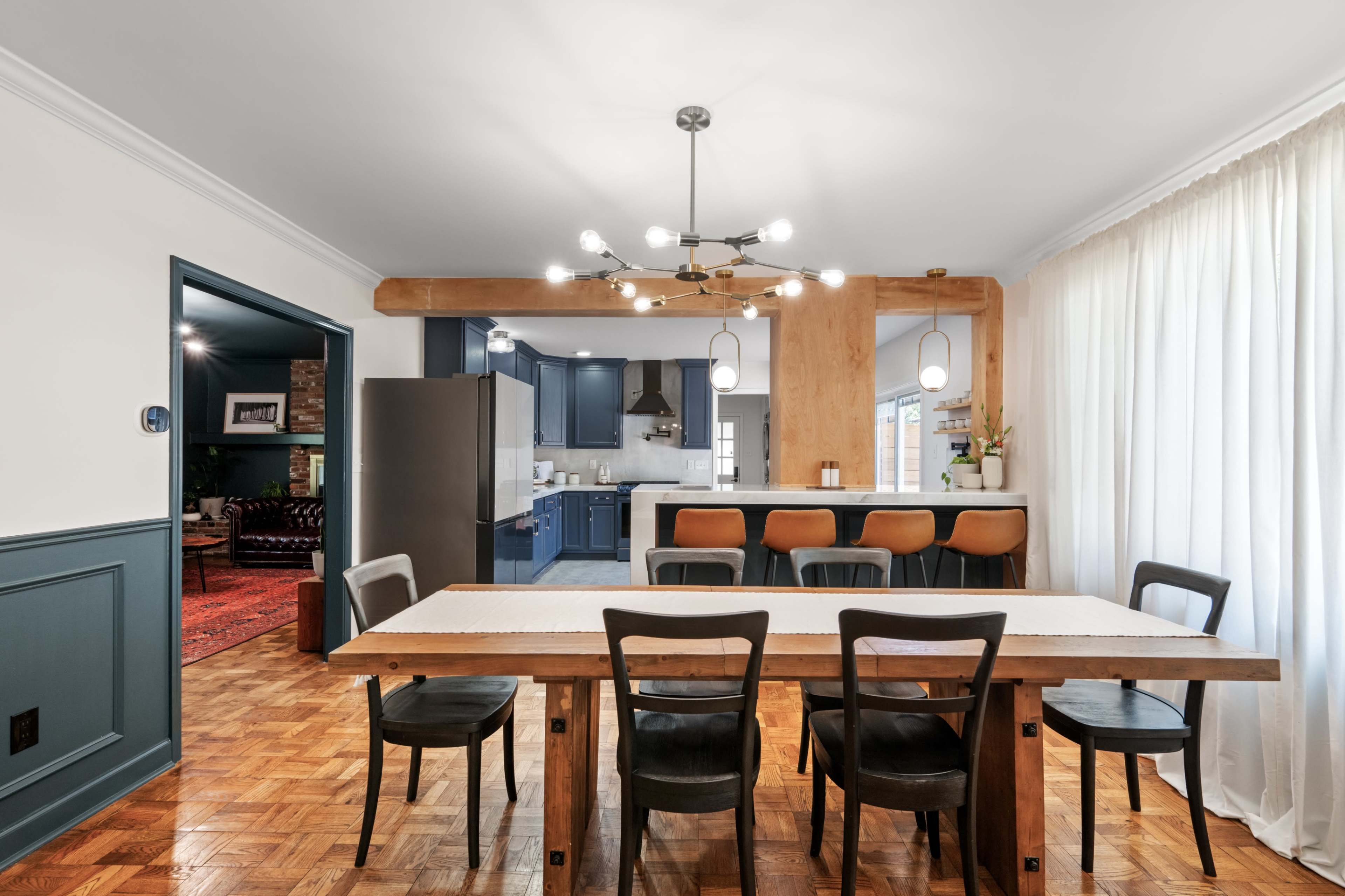 A spacious dining area features a wooden table surrounded by black chairs, with a contemporary light fixture overhead and a view of a kitchen in the background.