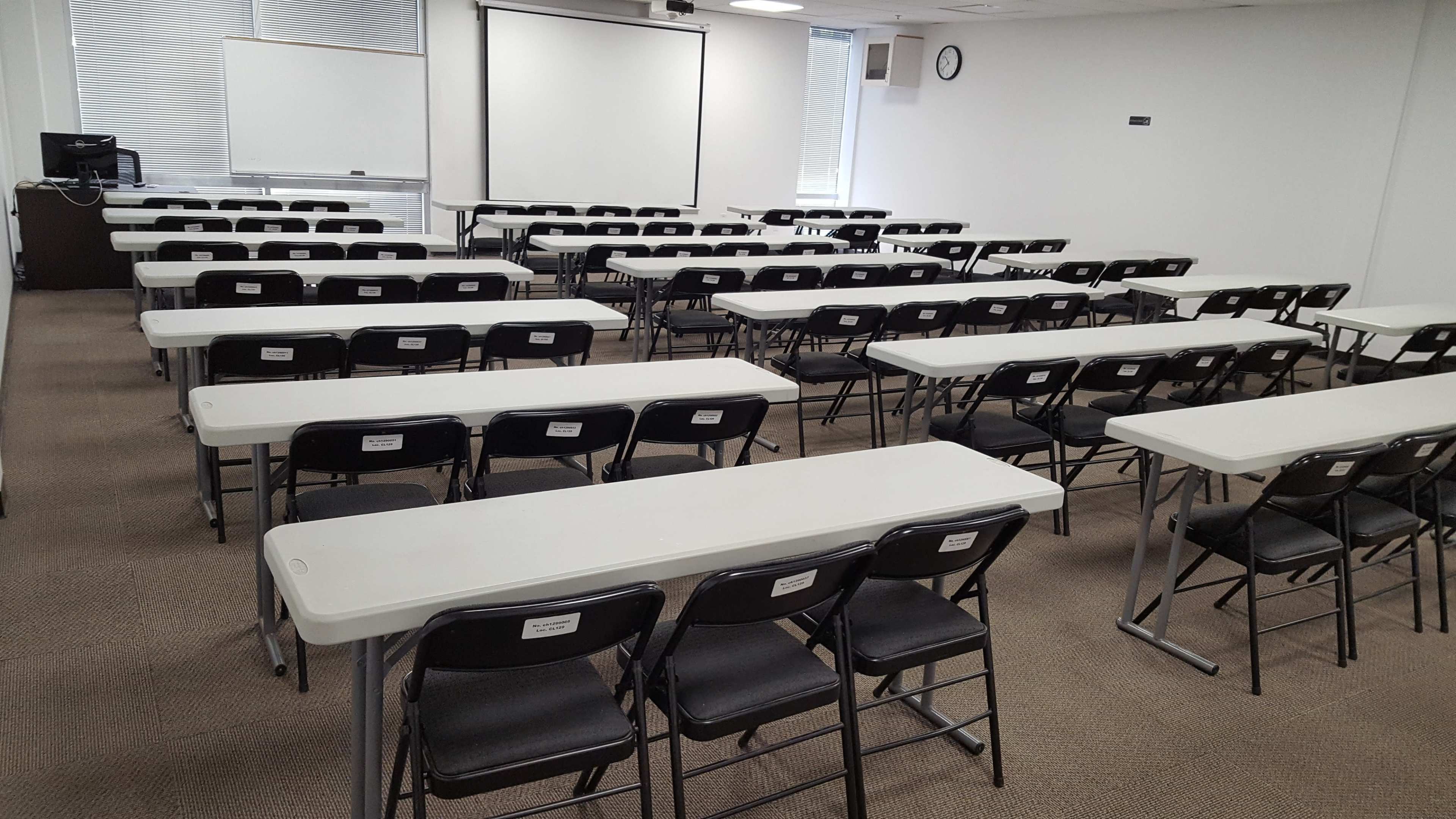 A classroom set up with multiple rows of tables and chairs, facing a white screen and a wall clock.
