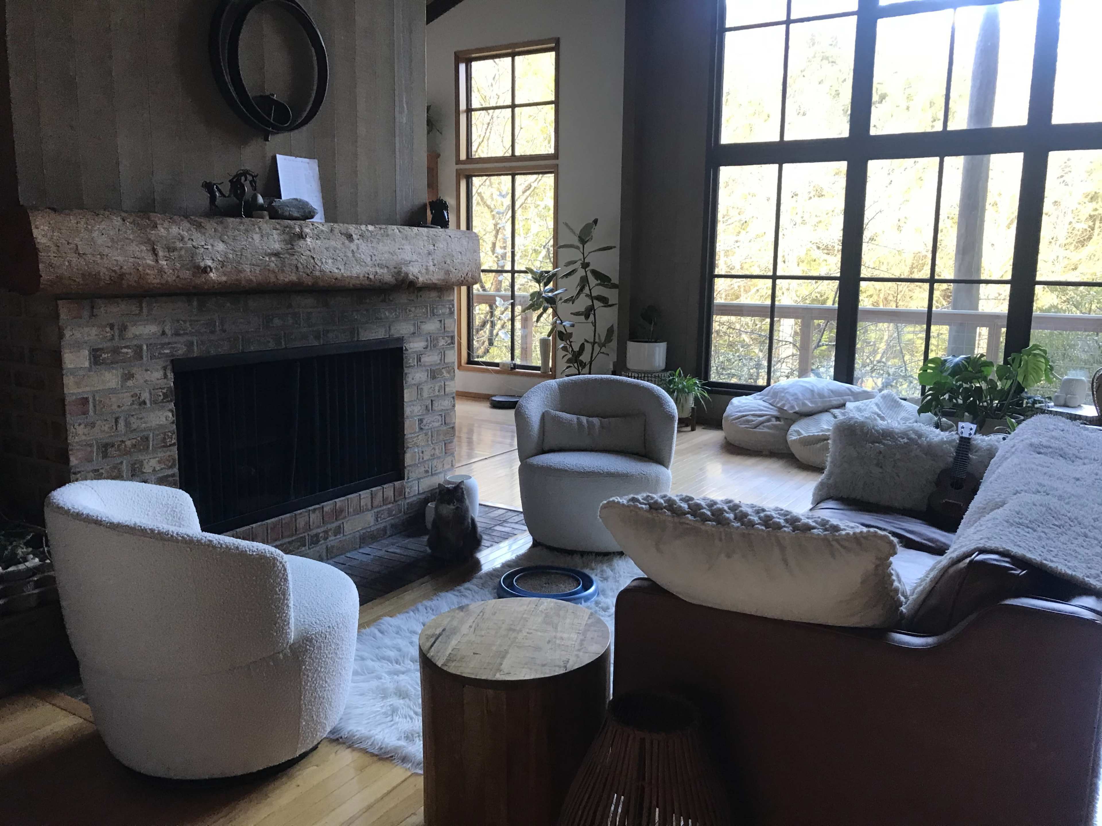 A spacious living room features a large fireplace, two round chairs, a wooden table, and large windows allowing natural light to fill the space.