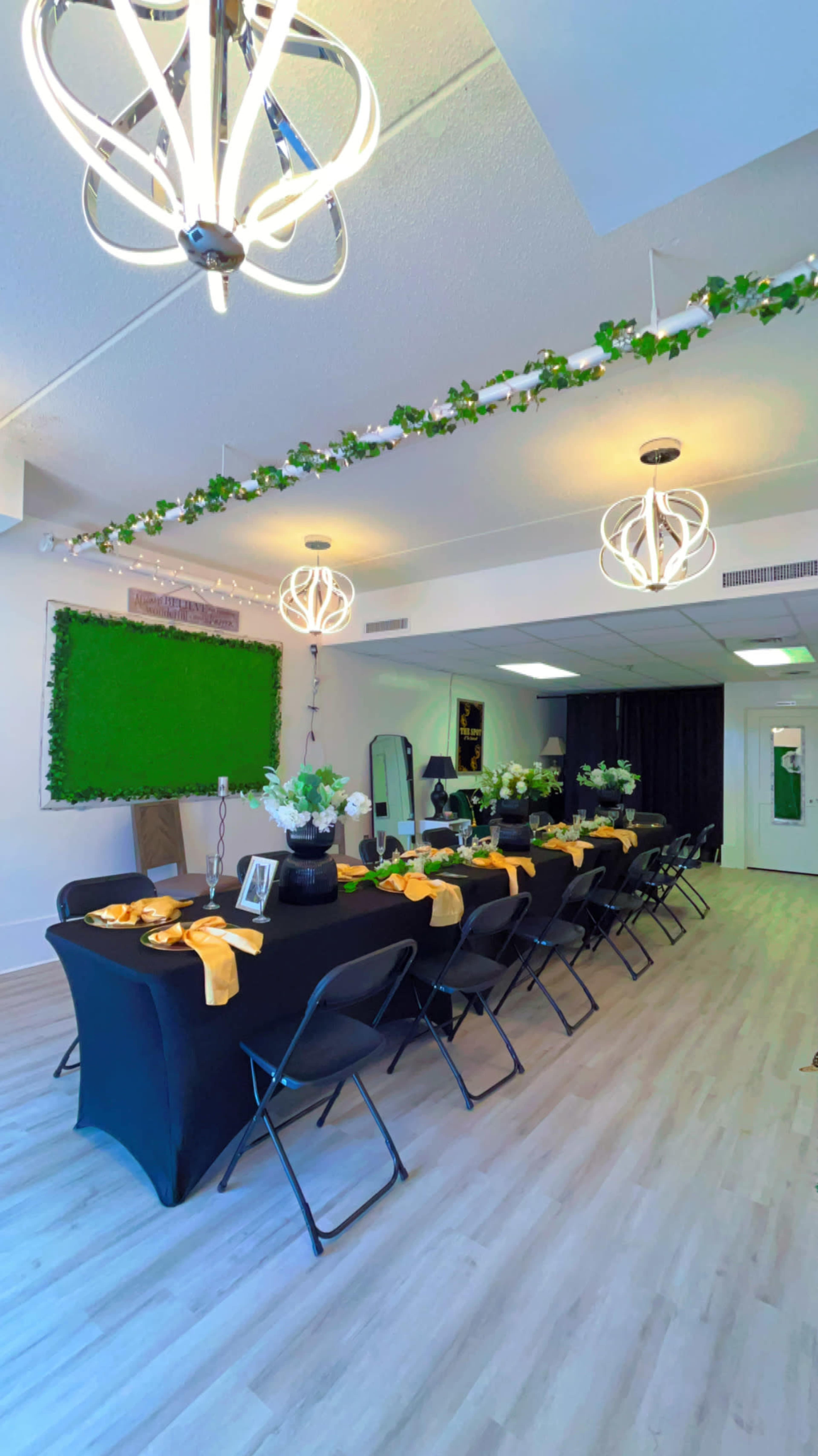 The image shows a decorated event space with a long table set for a gathering, featuring black tablecloths, yellow accents, and floral centerpieces, under modern light fixtures.