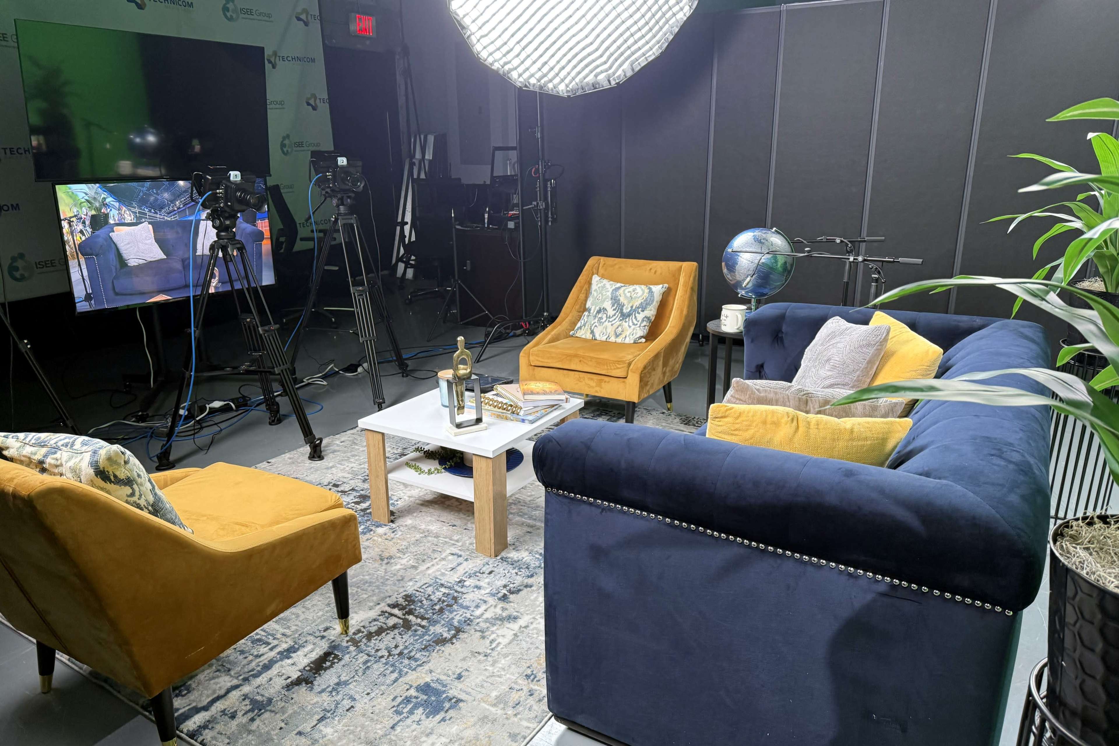 The image shows a well-lit studio setup with a navy blue couch, two yellow armchairs, a coffee table, and cameras positioned for filming.