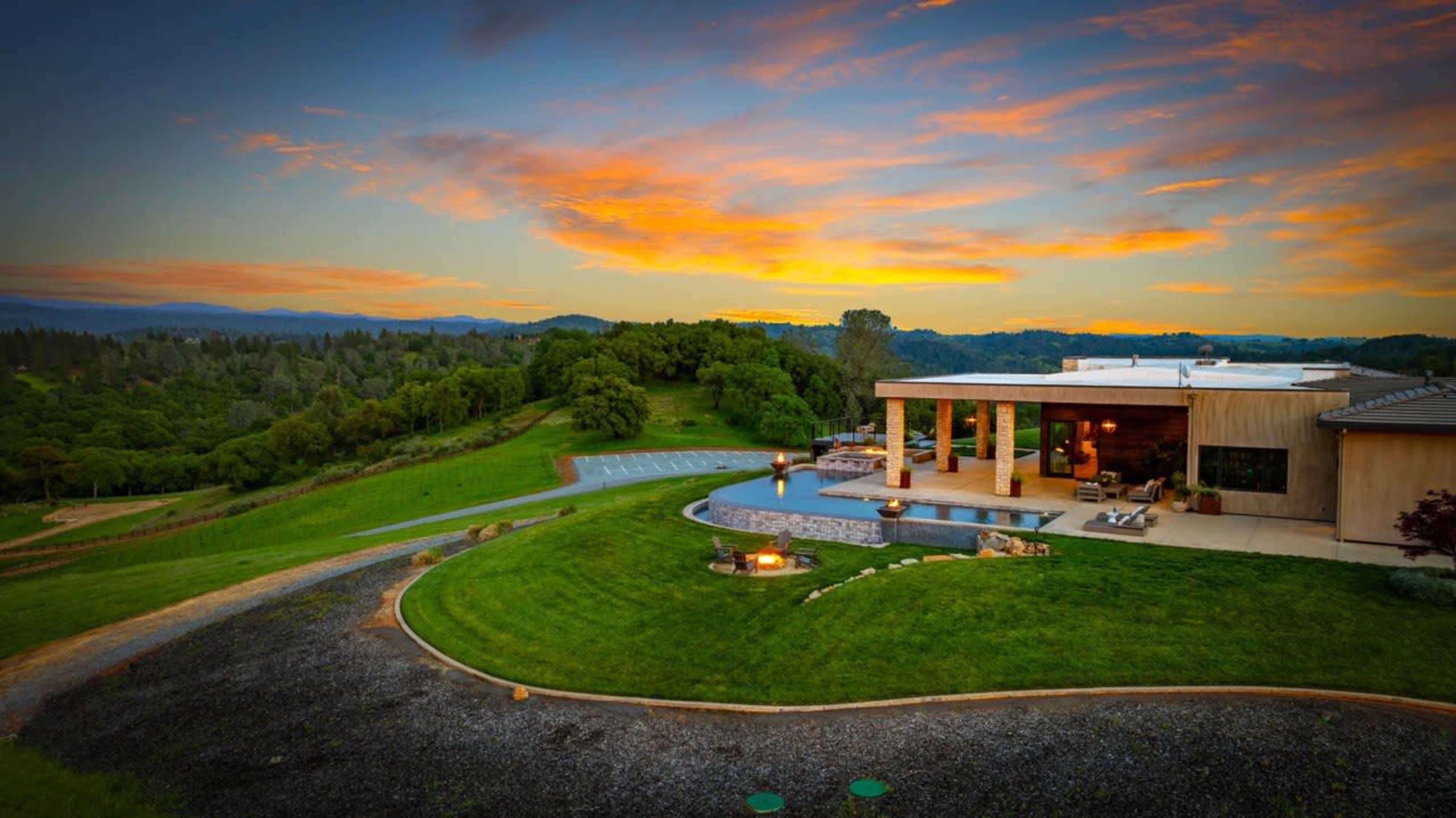 A modern house with a large pool and outdoor seating area sits on a hillside, surrounded by lush green landscape under a colorful sunset sky.