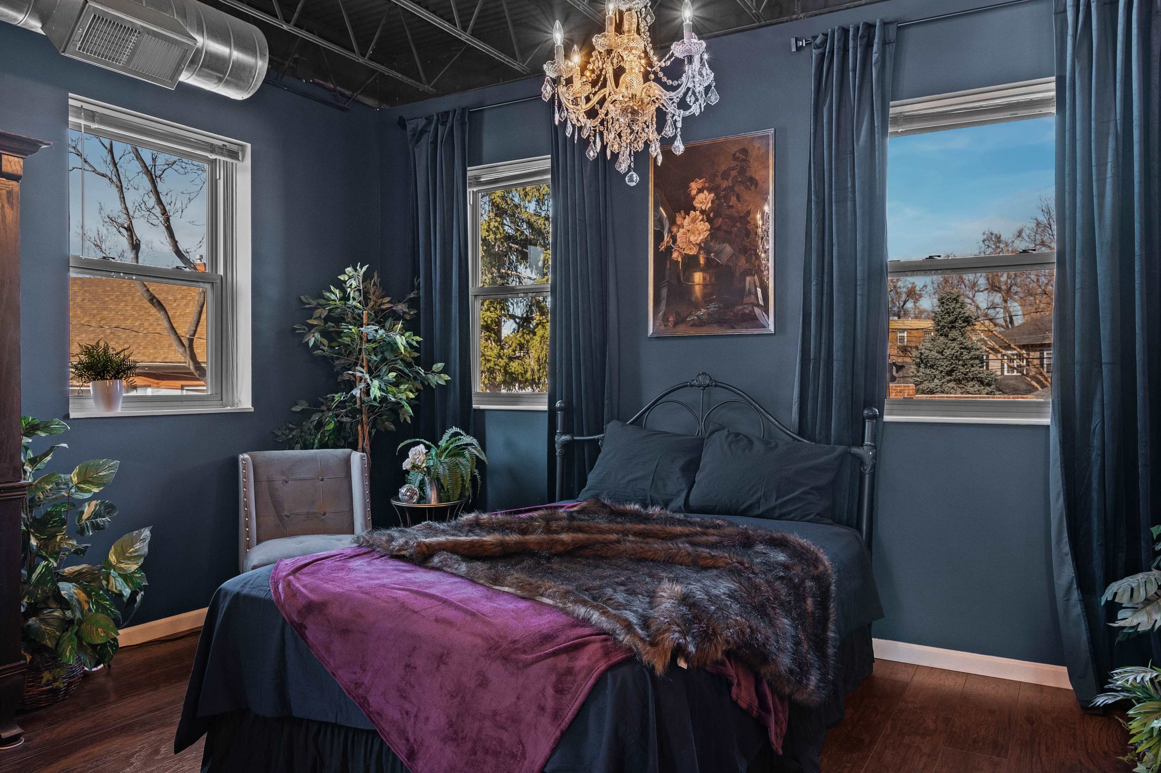 A bedroom featuring a bed with a purple blanket, a chandelier above, and large windows with views of trees outside.
