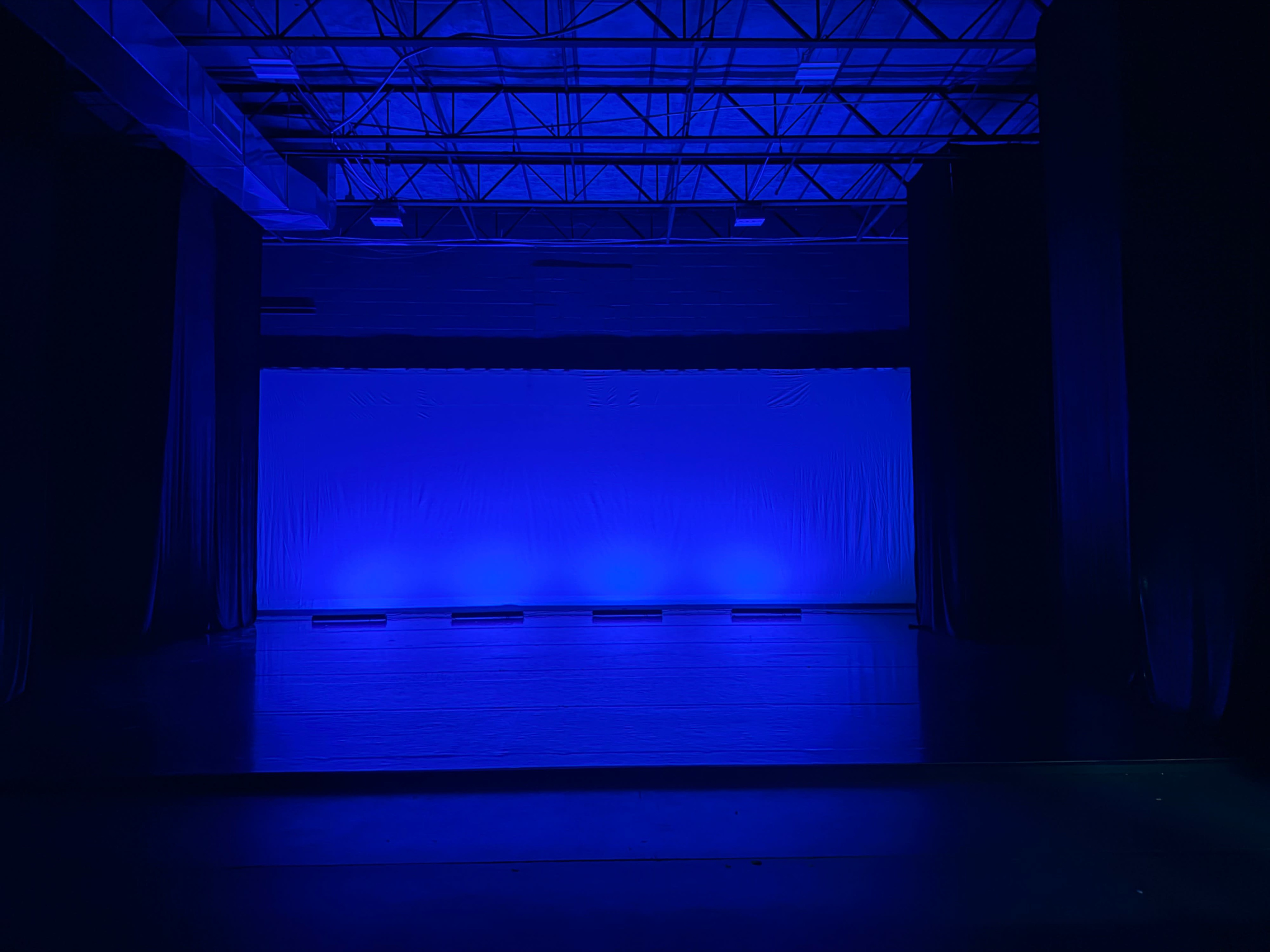 The image shows an empty stage illuminated by blue lights, with a plain backdrop and black curtains on either side.