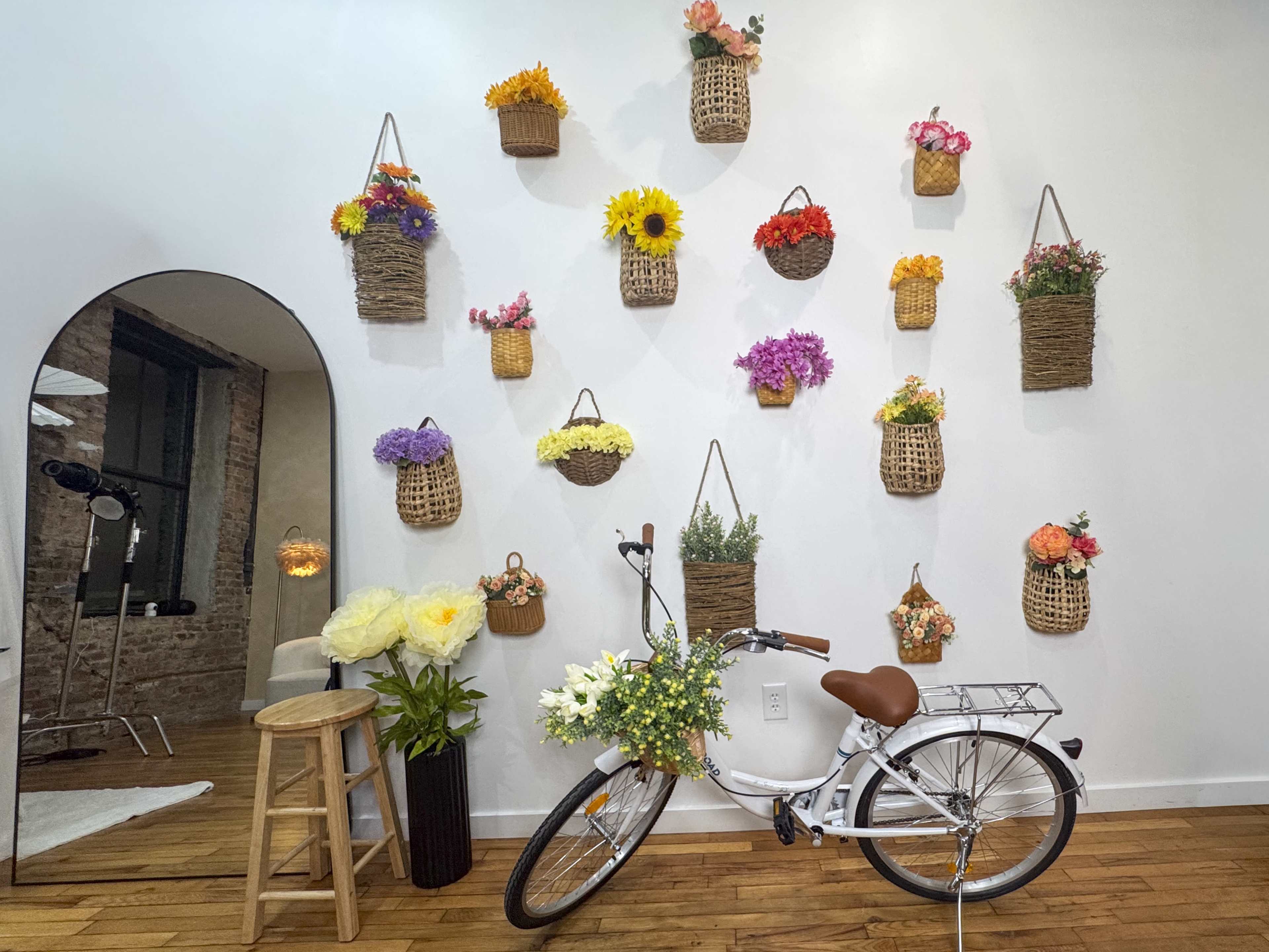 A white bicycle adorned with flowers is positioned beside a wall decorated with woven baskets filled with colorful plants and blooms.