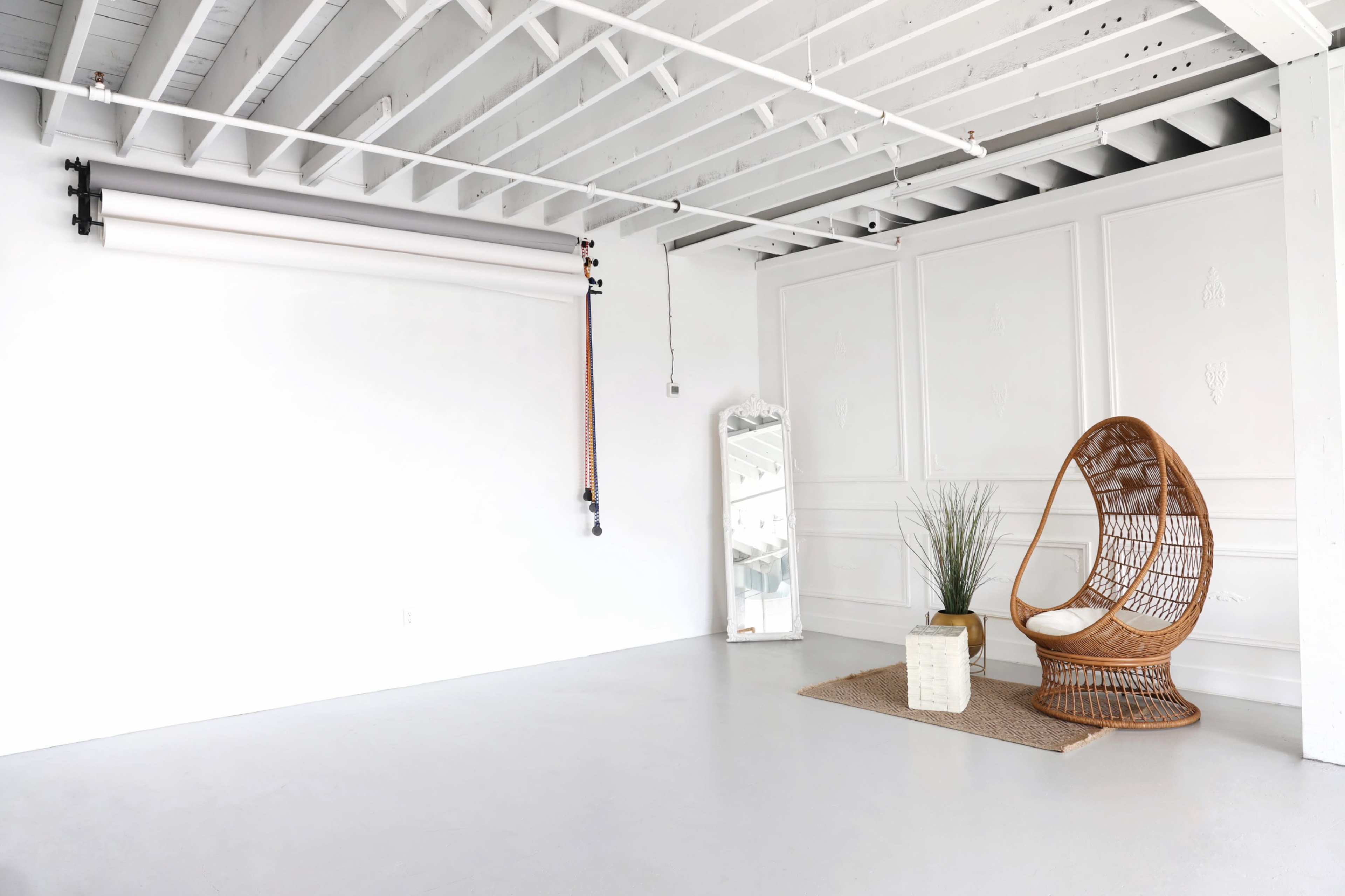 A minimalist room featuring a rattan chair, a floor mirror, a plant in a beige pot, and a rug on a concrete floor under a ceiling with white beams.