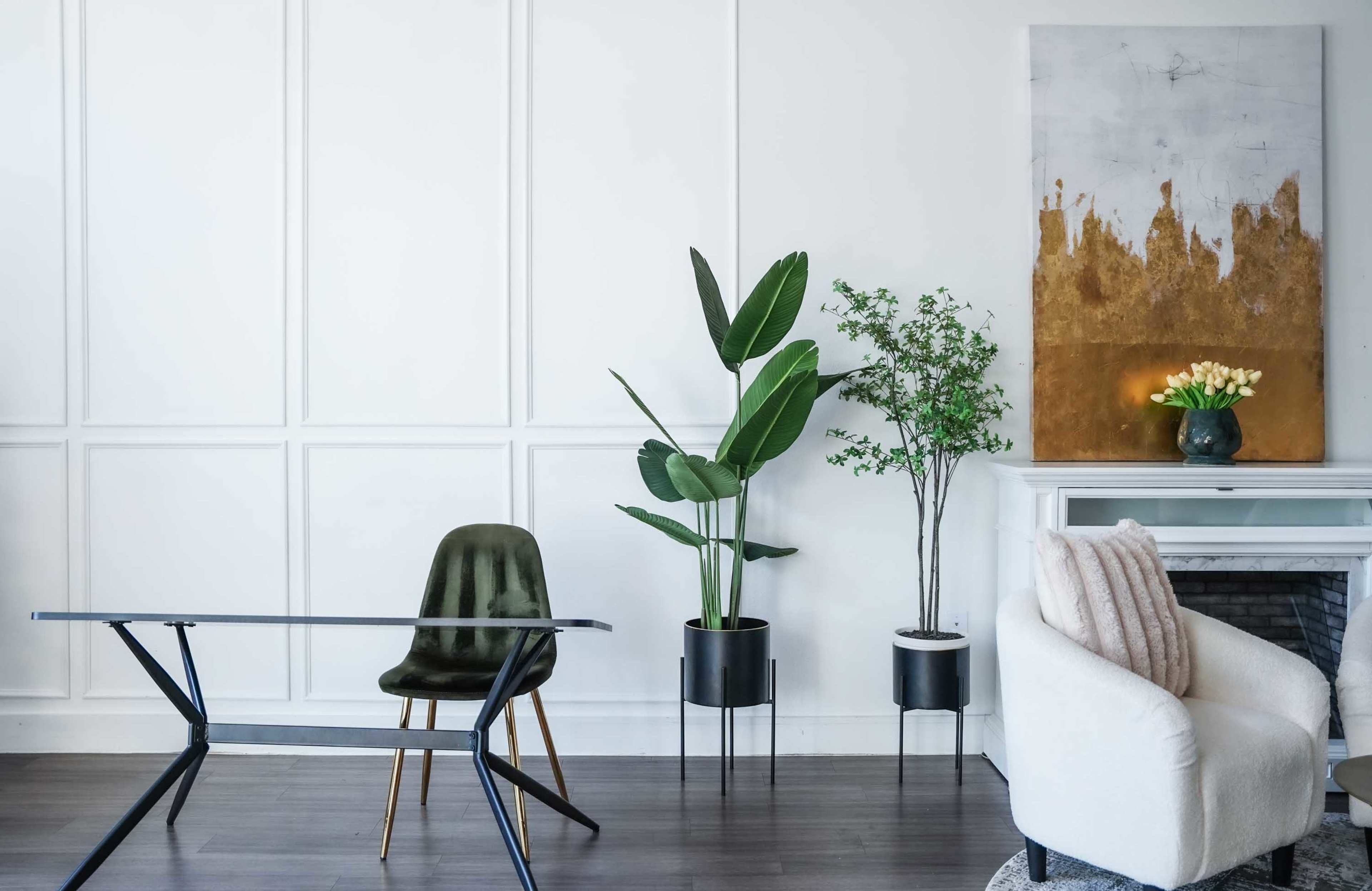 The image shows a modern interior with a glass desk, a green upholstered chair, potted plants, and a white armchair beside a fireplace.